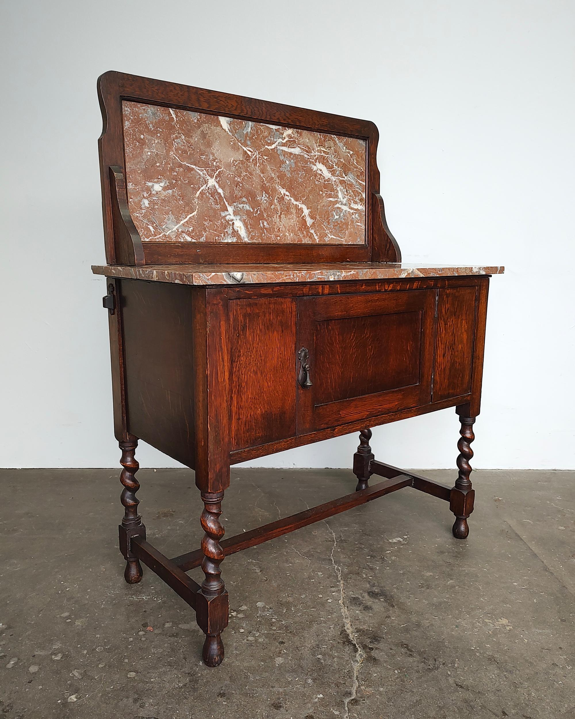 Early 20th-century oak wood wash stand cabinet with original pink/grey stone top and backsplash, and beautiful carved barley twist legs. One cabinet door with patinaed brass drop pull hardware. Inside opens to large storage compartment. Overall