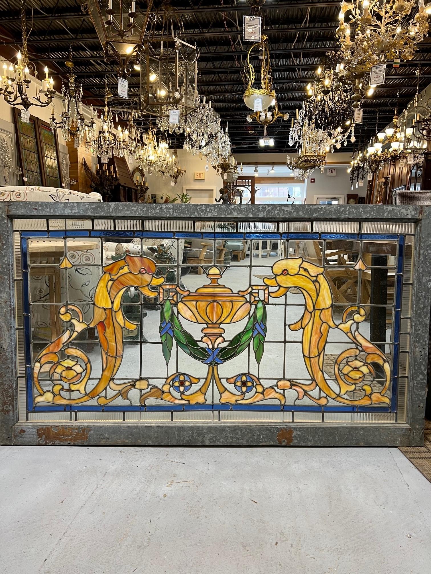 Early 20th century antique stained glass window with Victorian dolphins on either side of a draped covered urn. Its a nice combination of stained glass and clear glass panels in a galvanized wood frame. I have two identical windows available for