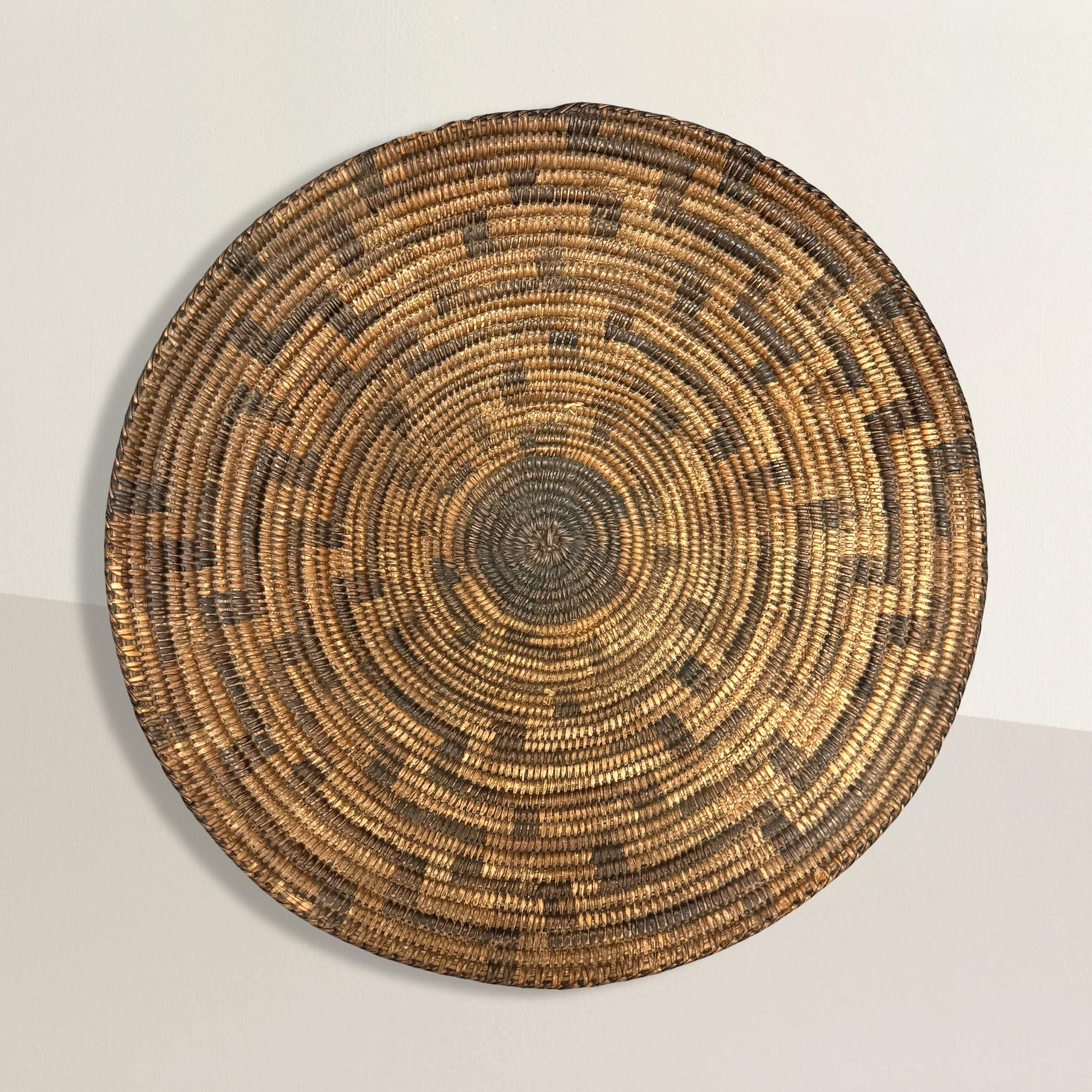 This early 20th-century Apache tray is a stunning example of traditional basketry, hand-woven with precision from willow and devil’s claw fibers. The design features a striking counter-clockwise fretted spiral pattern, radiating outward from the
