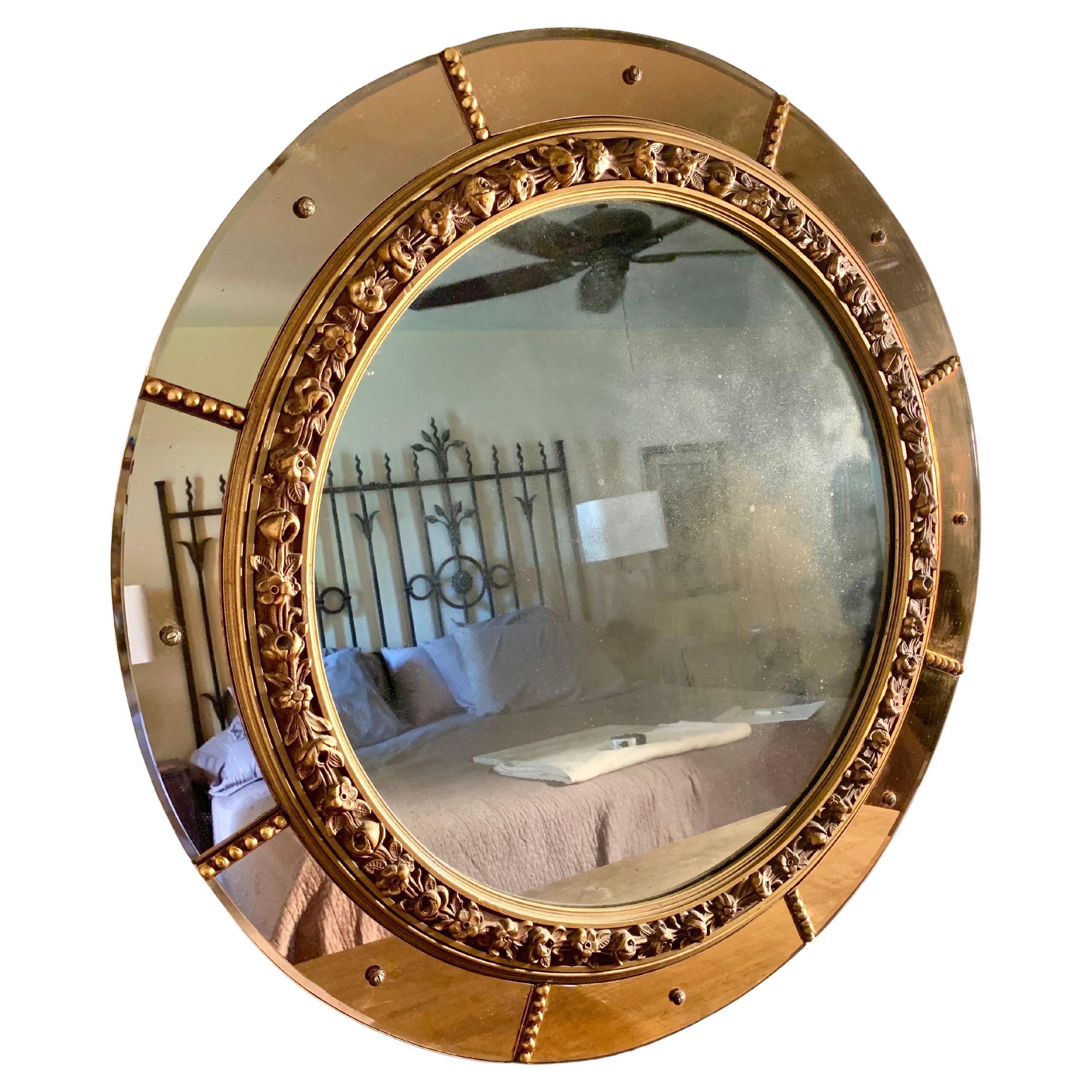 A true testament to the Art Deco Era, this Early 20th Century Circular Wall Mirror presents a round mirror surrounded by a gold floral design. The outer edge of the mirror is surrounded by eight pink or rose colored mirrored glass panels that have