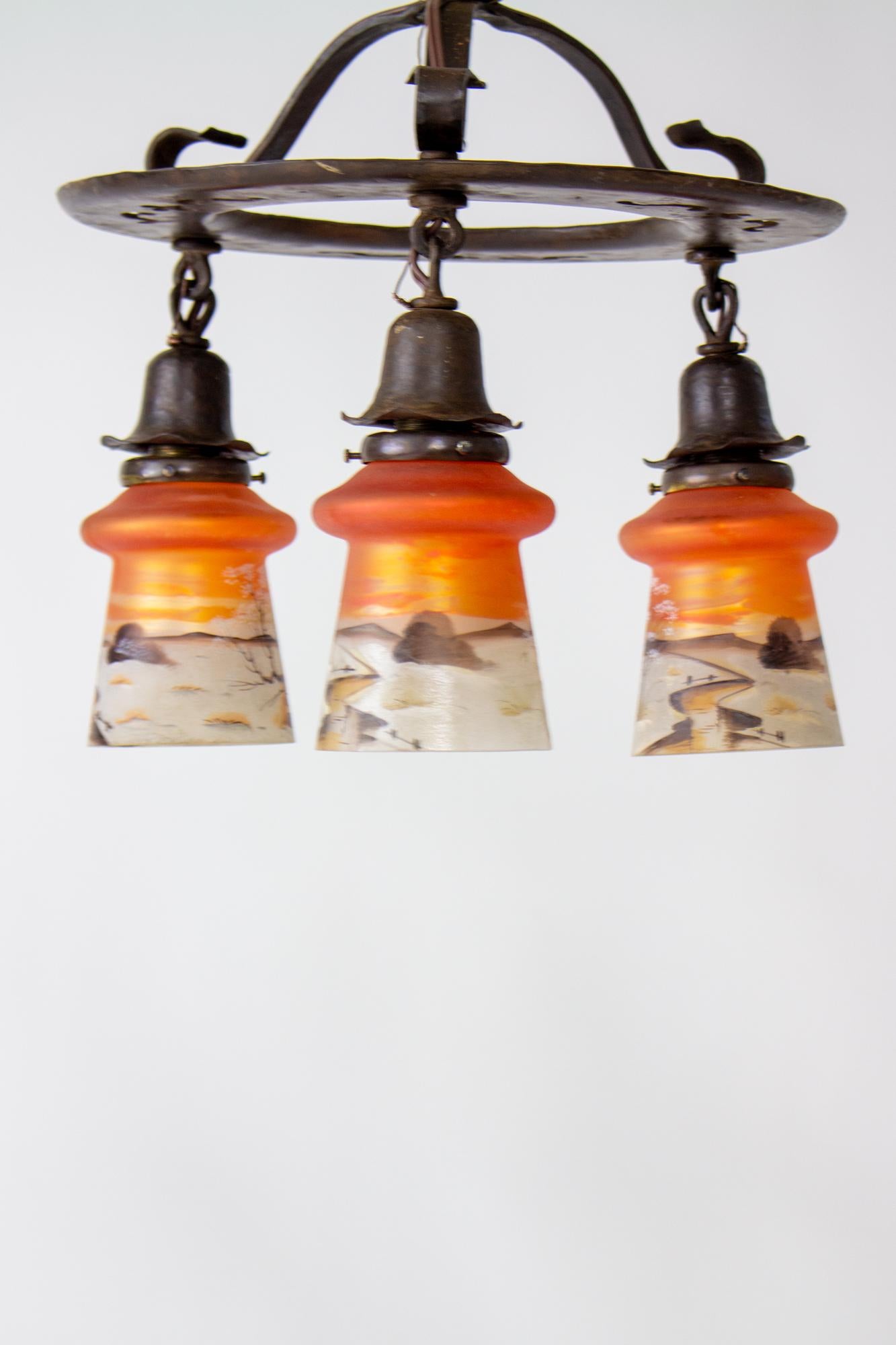 Early 20th century arts and crafts chandelier with orange painted glass. Wrought iron and brass fixture with three down hanging lights. The glass is orange with a hand painted scene. Iron has been cleaned and waxed, rewired, and glass in very good
