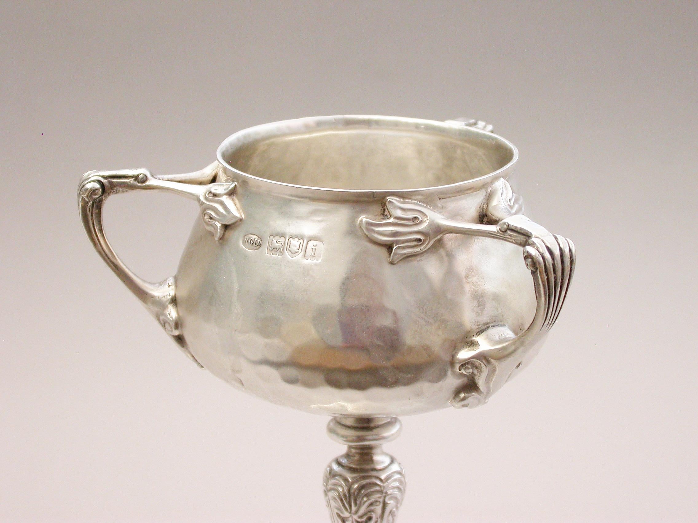 An early 20th century Arts & Crafts style hammered silver cup, with three applied handles and chased stem, of a good gauge and weight. A later (1960) engraved inscription to the base.

By H Greaves Ltd, London, 1904.

In good condition with no