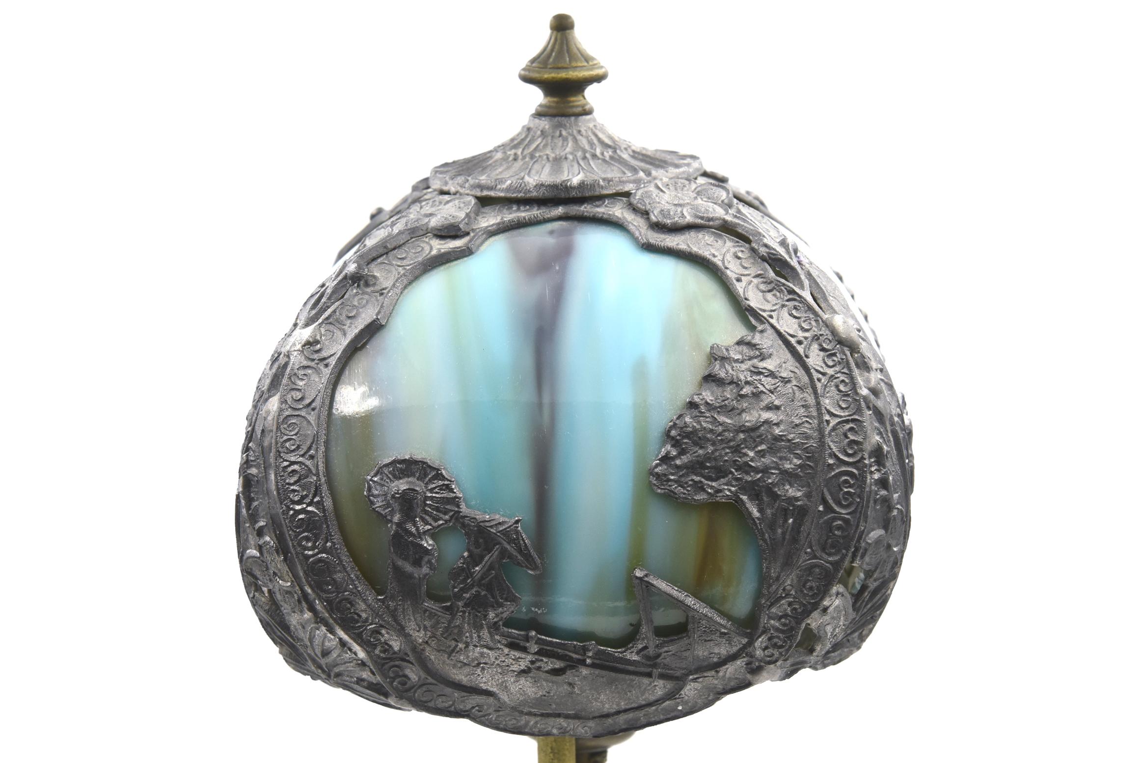 Early 20th century metal table, parlor or boudoir lamp featuring an Oriental motif lined with four bent slag glass blue and green panels. The scene illustrates two woman talking under umbrellas with a small dog running ahead near a tree. Along the