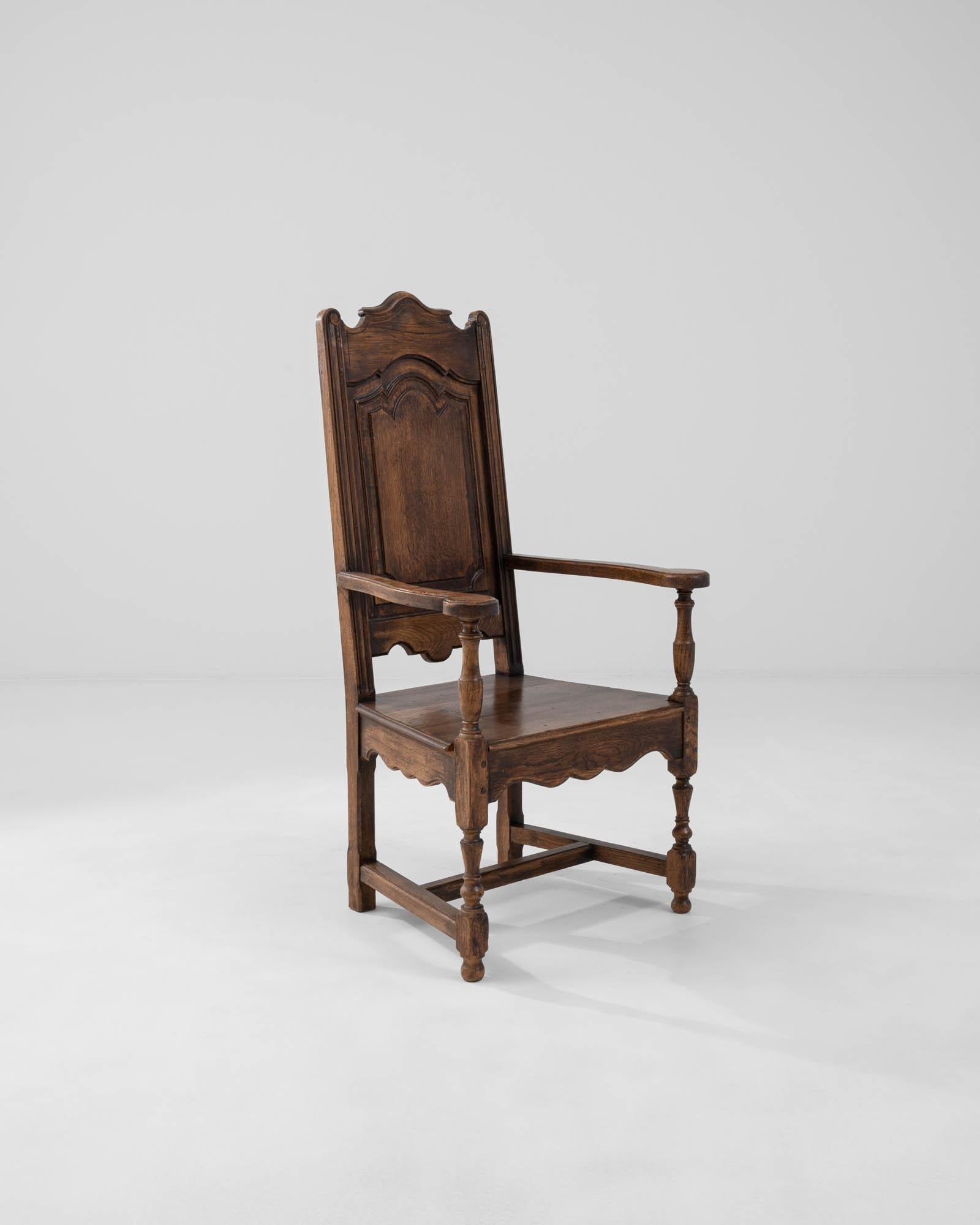 A wooden armchair created in early 20th century Belgium. High-backed and dignified, this sumptuous armchair radiates an aura of royal authority. Lavishly sculpted aprons, lathed legs, and a paneled back demonstrate a flair for the dramatic and