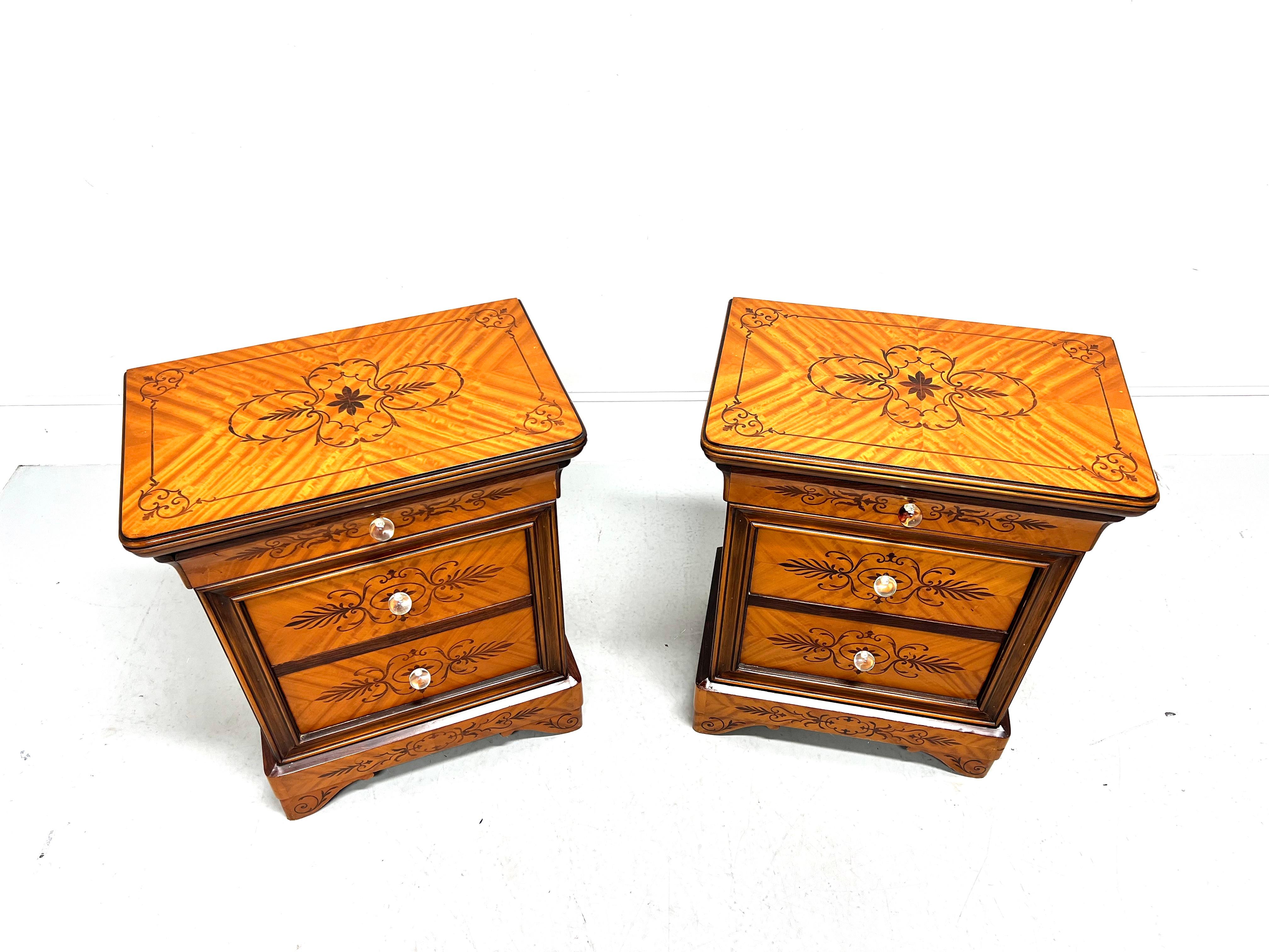 A pair of Biedermeier style nightstands, unbranded. Nutwood with decorative marquetry designs, marquetry to top with ogee edge & rounded corners, crystal knob hardware, and bracket feet. Features three drawers of dovetail construction. Likely made