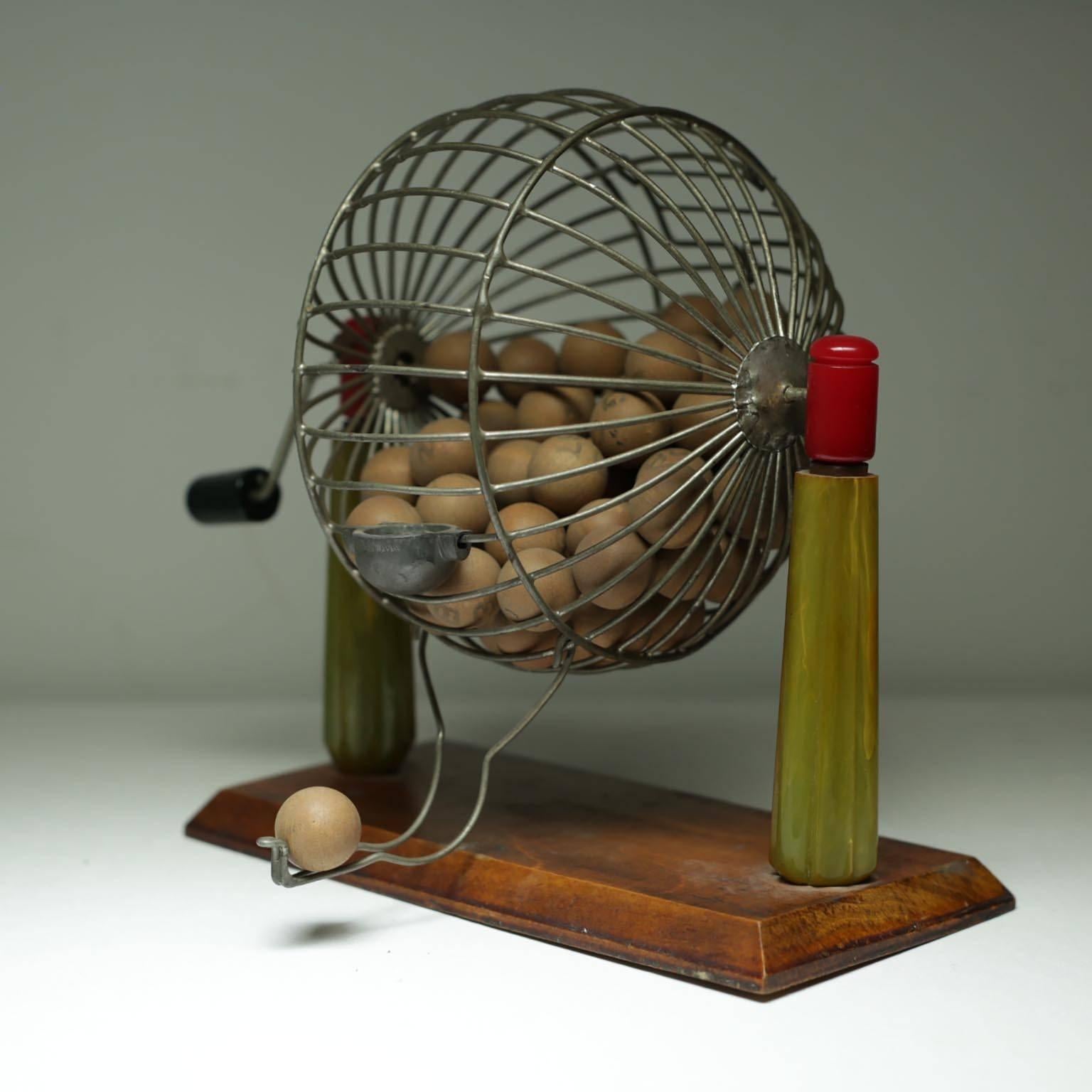 Wire bingo cage with beautiful green and red Bakelite supports on a wooden base. Comes with a stack of original bingo playing cards and original chips in a caddy. All of the balls are numbered and lettered.
  