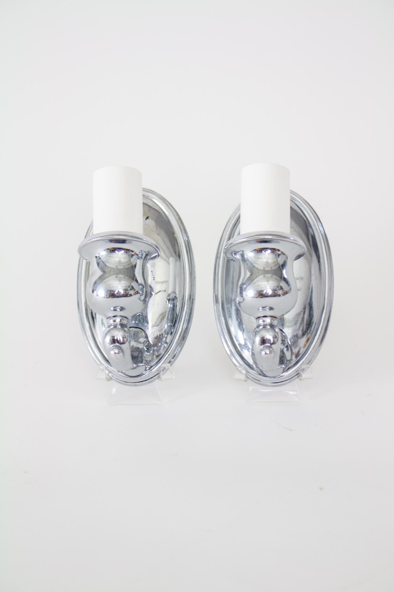 Bradley and Hubbard chrome sconces - a pair. Small sconces, marked with B&H on the backplate. US c. 1910. Oval backplate and small single arms with a curvy candle cup. White candle covers. Finish is in good condition, some chipping to the chrome