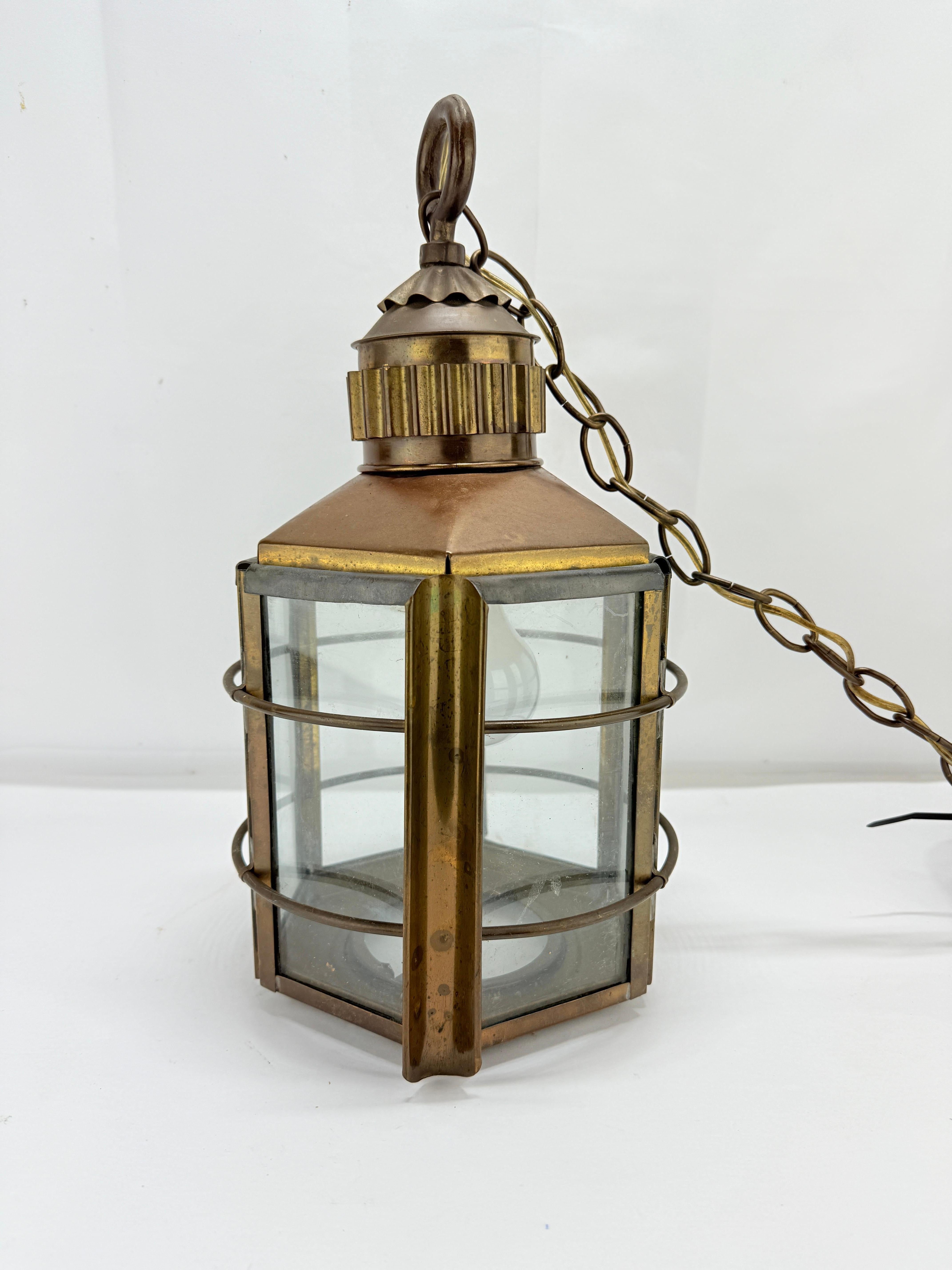 This antique brass and glass hanging lantern exemplifies the robust craftsmanship and utilitarian elegance characteristic of early 20th-century maritime and industrial lighting. Its hexagonal framework, constructed from aged brass with a rich,