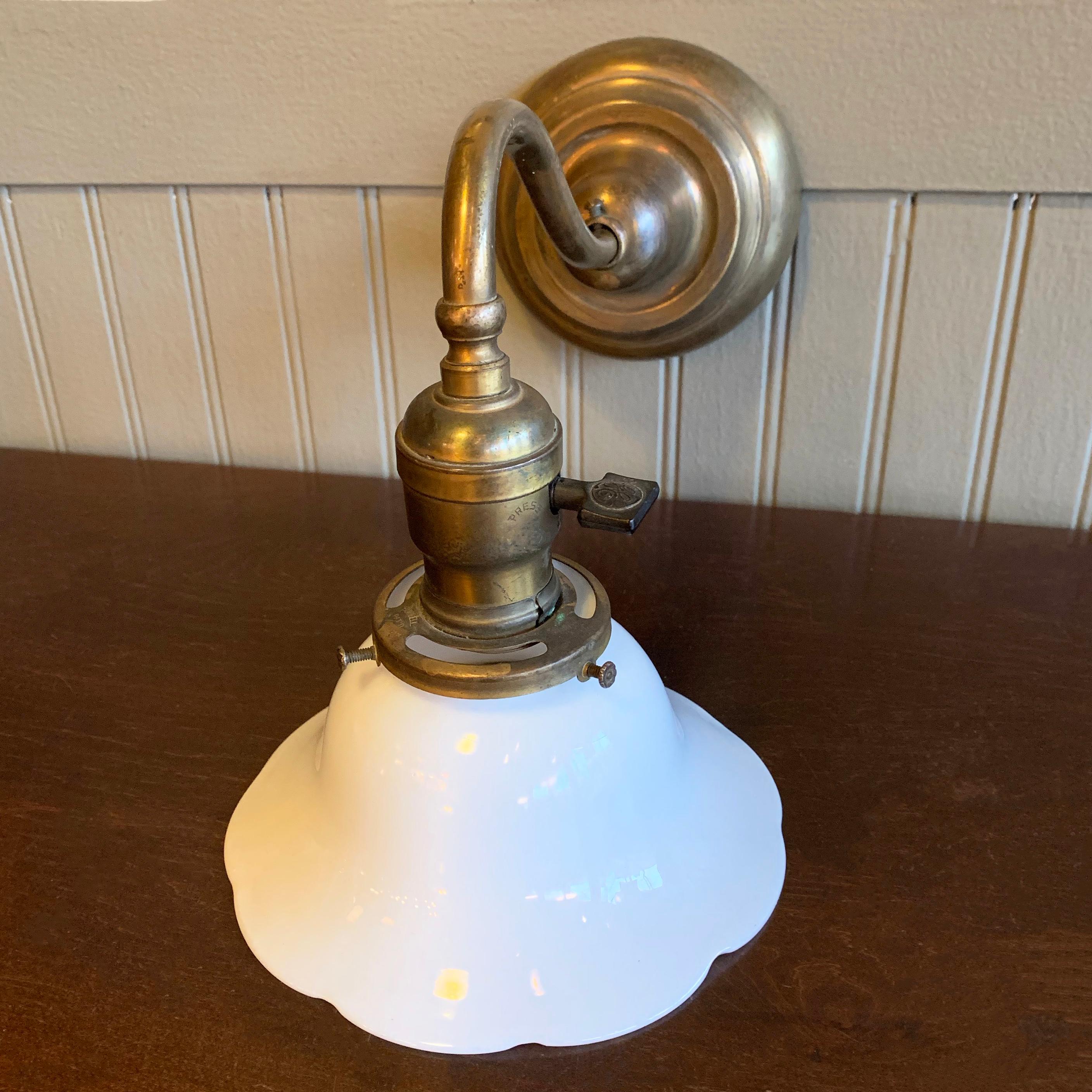 Early 20th century wall sconce lamp features a curved brass stem with ruffled edge, milk glass shade. The sconce is wired to accept up to a 100 watt bulb. The backplate measures 4.5 inches diameter.