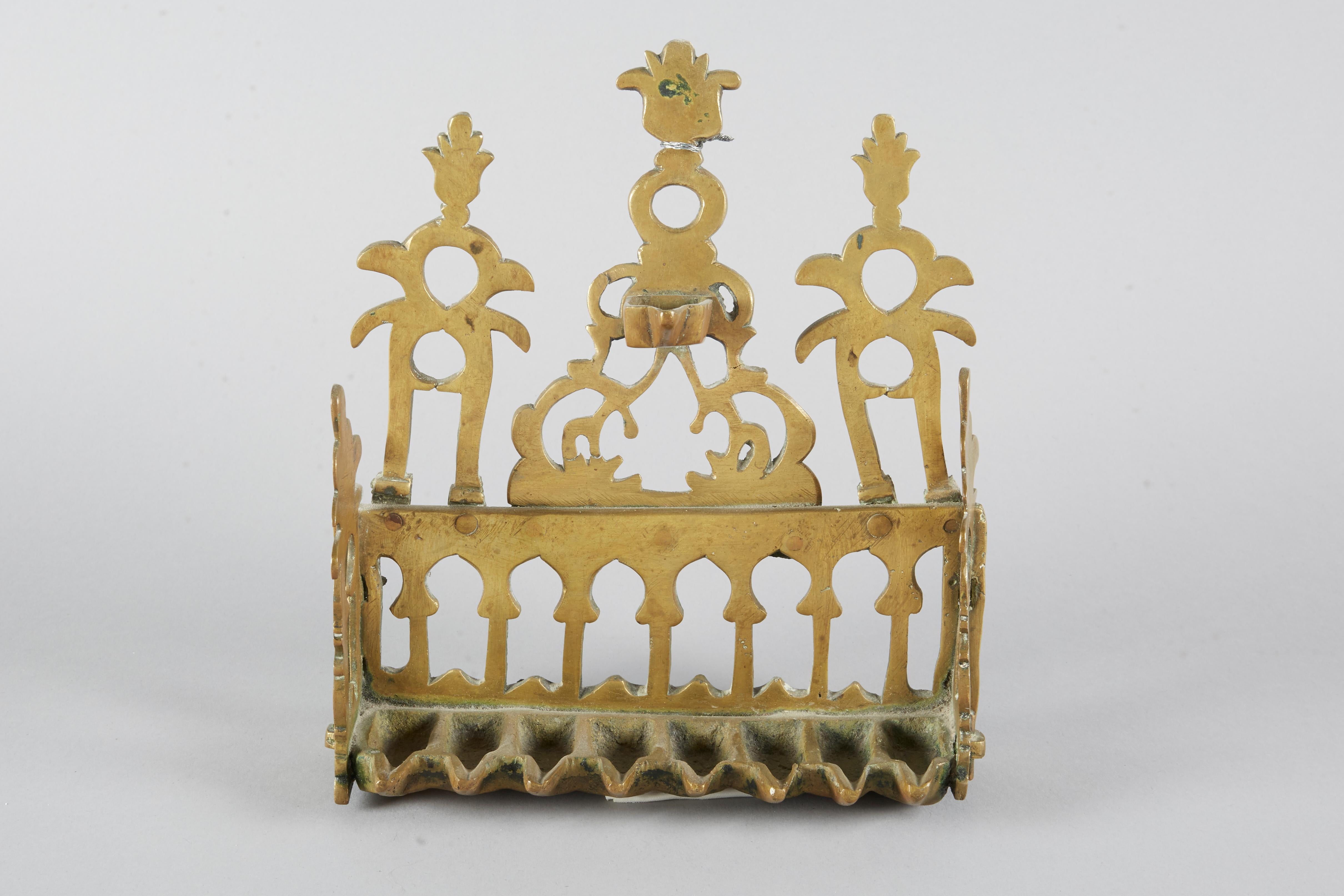 The hanging back-walled Hanukkah Lamp is made of brass and consists of a row of oil containers, a back and sides wall, and a hanger. A row of eight rectangular spouted oil containers is attached to the back and side walls. The pointed arch openwork