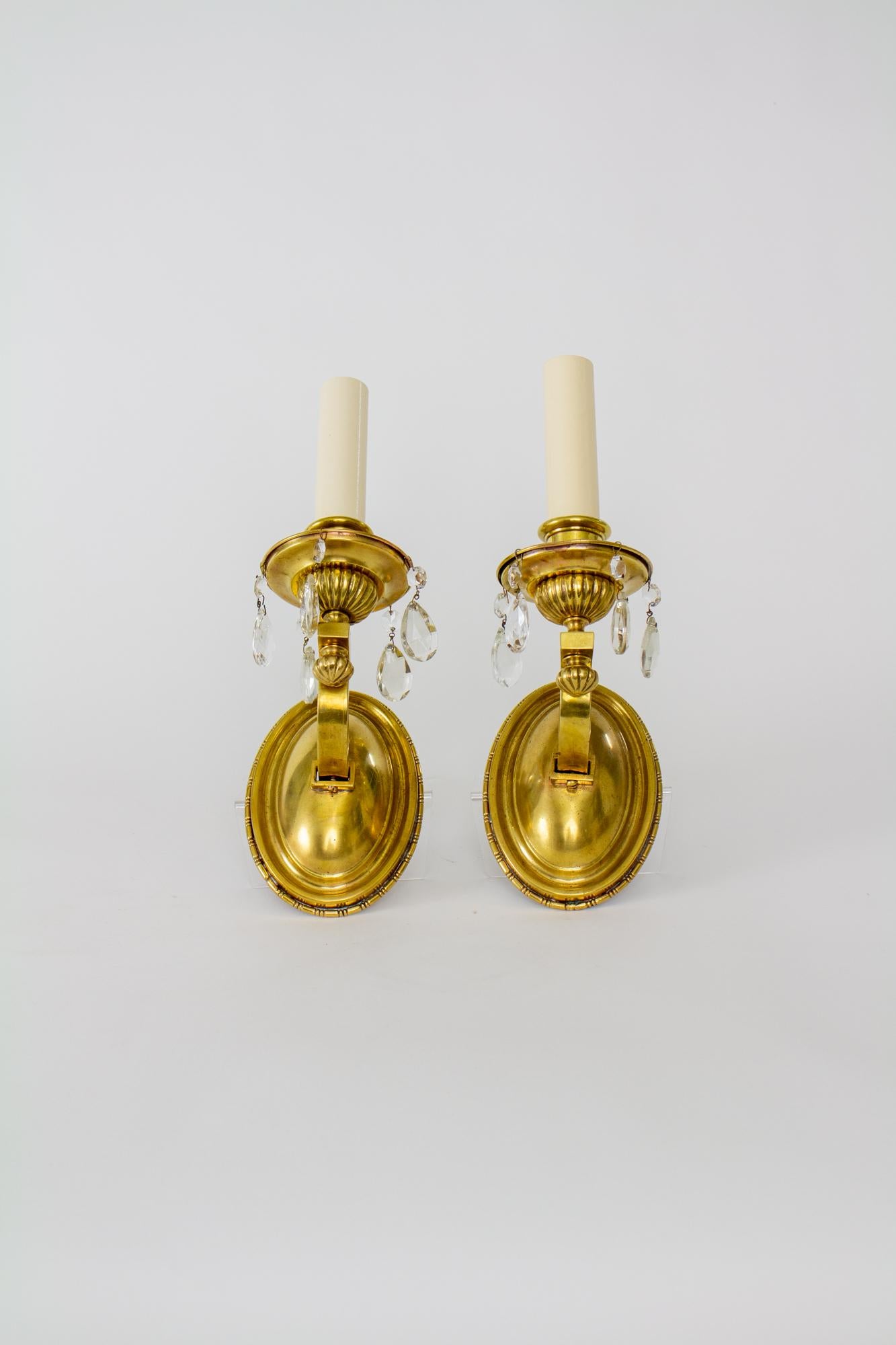 Early 20th Century brass sconces with crystals - a pair. Oval backplate with curved arms made of rectangular tubing. Ribbed decorative elements on rim of backplate, finial, and bottom of bobeche. Teardrop crystals hang from the bobeche. White