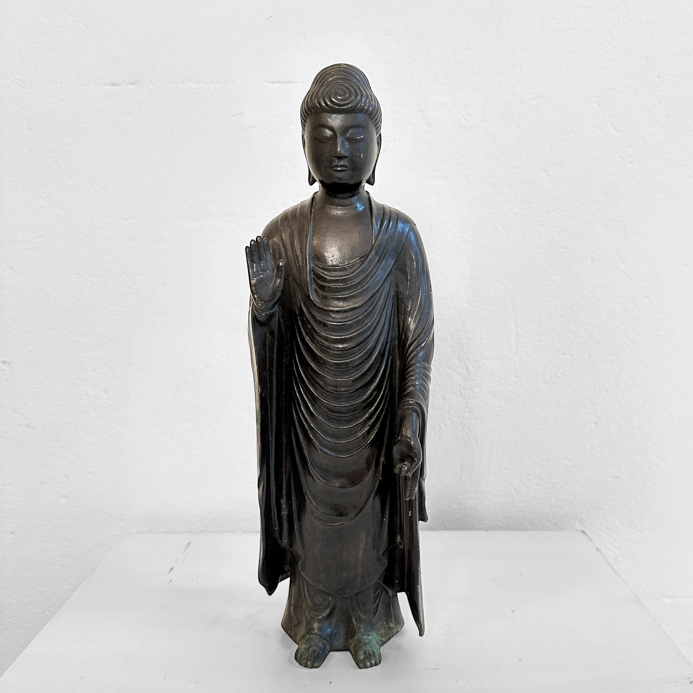 This captivating bronze Buddha sculpture, originating from Thailand around 1920, embodies the spiritual serenity of the early 20th century. The standing figure is meticulously crafted, showcasing exceptional detail in the serene and well-carved