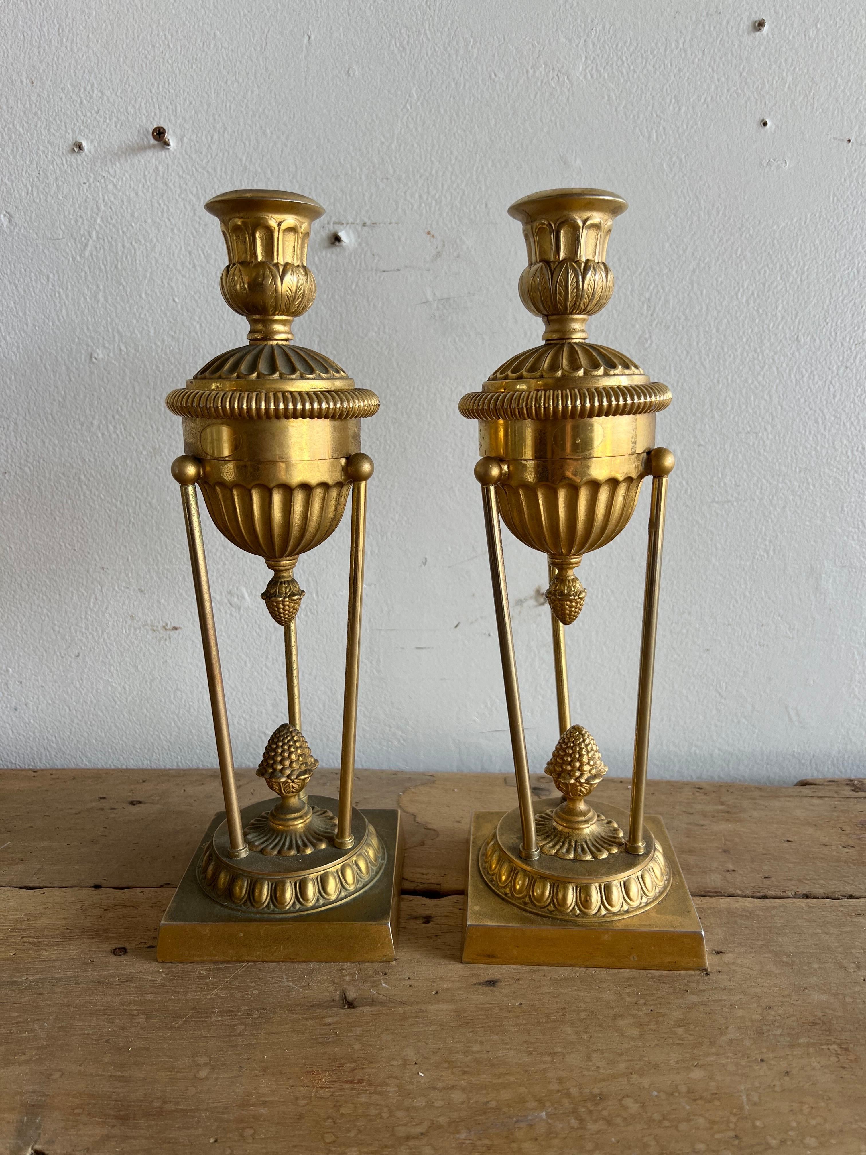 Questi portacandele sono una coppia di pezzi in bronzo in stile classico.
Presentano un design con una parte superiore scanalata e una forma arrotondata simile a un'urna al centro.
I supporti sono sostenuti da tre sottili aste collegate a una base