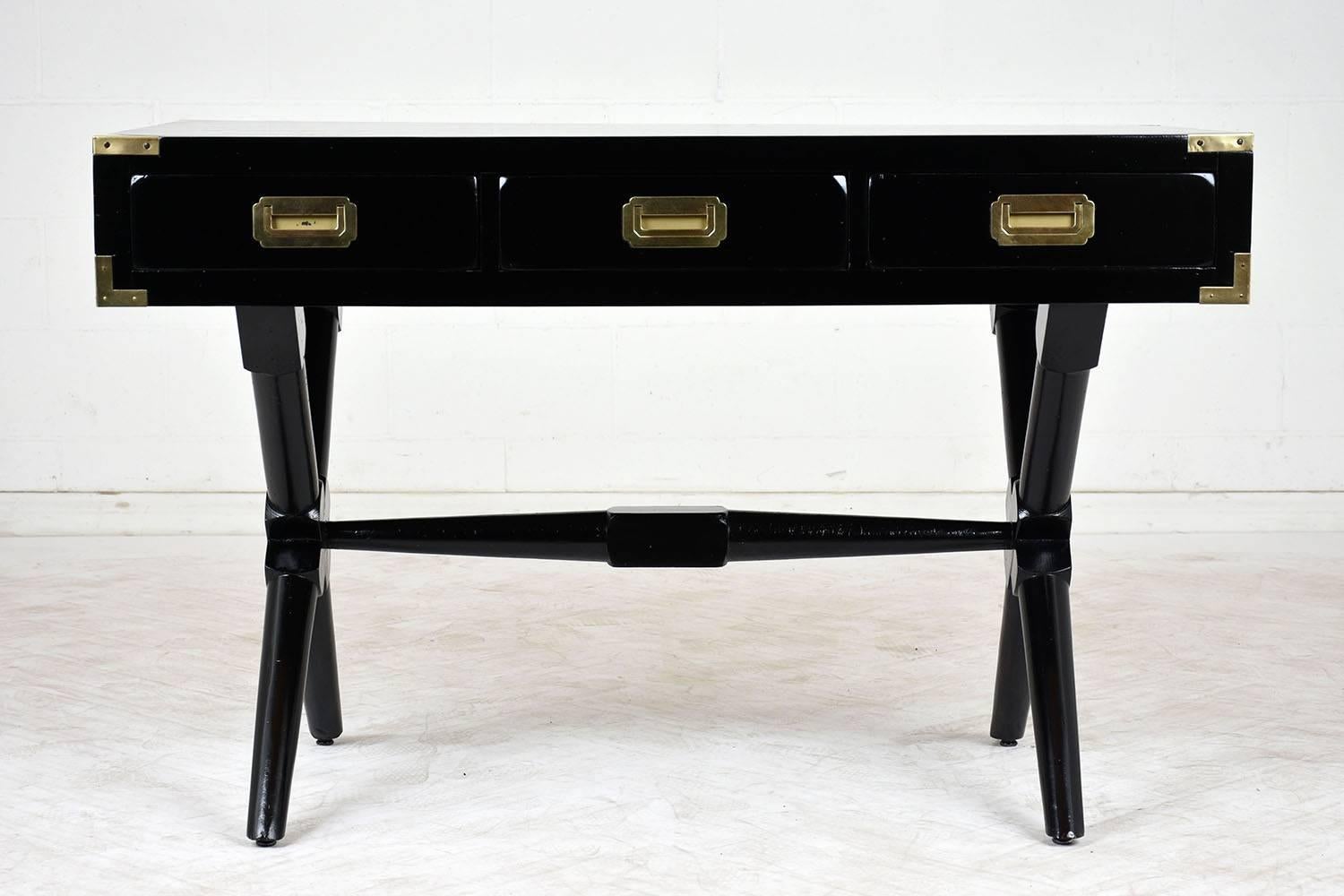 This completely restored 1930s Campaign-style writing desk is made of solid wood with an ebonized and lacquered finish. The desk has three-drawer with brass drawer pulls and brass accents on the desk corners. The X-shaped stretched legs are very