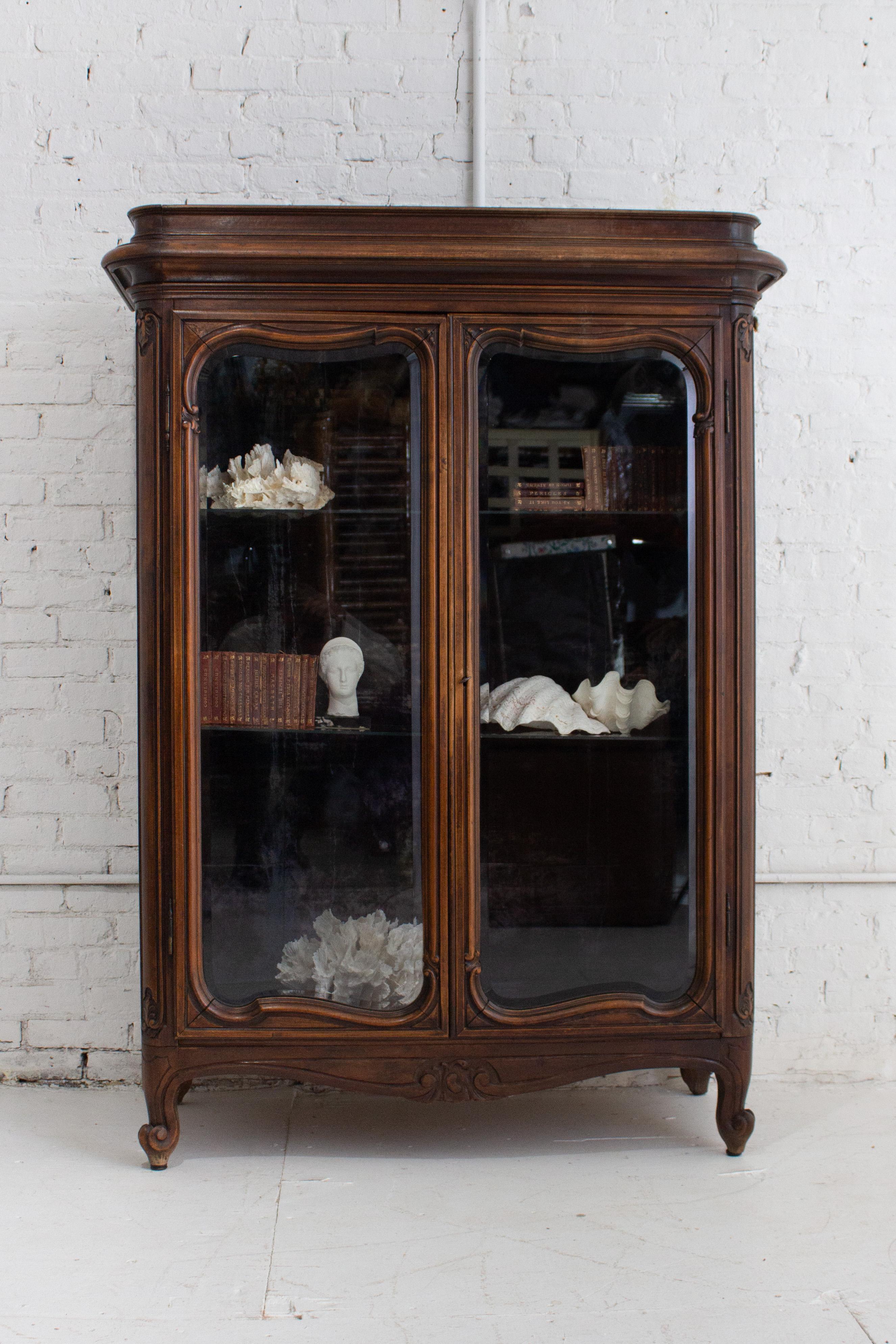 An early 20th century curio cabinet of carved dark walnut wood and beveled glass doors. Blue crushed velvet interior and glass shelving. Interior is presented as found, exhibiting a natural sun faded patina and signs of use from previous owners.