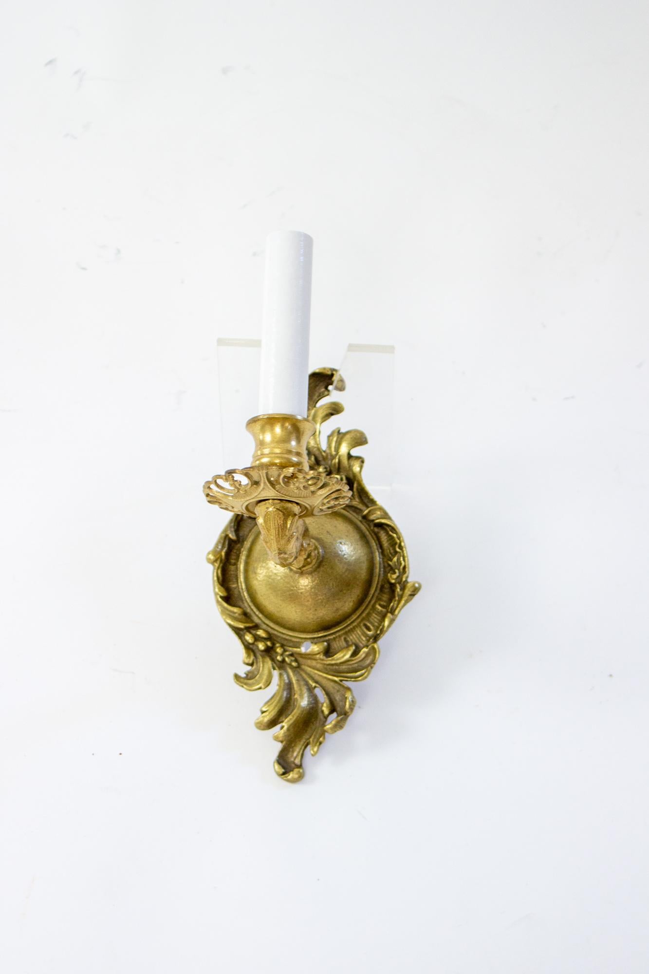 Cast brass sconces from the early 20th Century. Single arm each, with candelabra socket. Backplate is a dome surrounded by swirling leaves. Ornate cast arms, and bobeches with decorative cut outs, lightening up the sconces for a more delicate look.