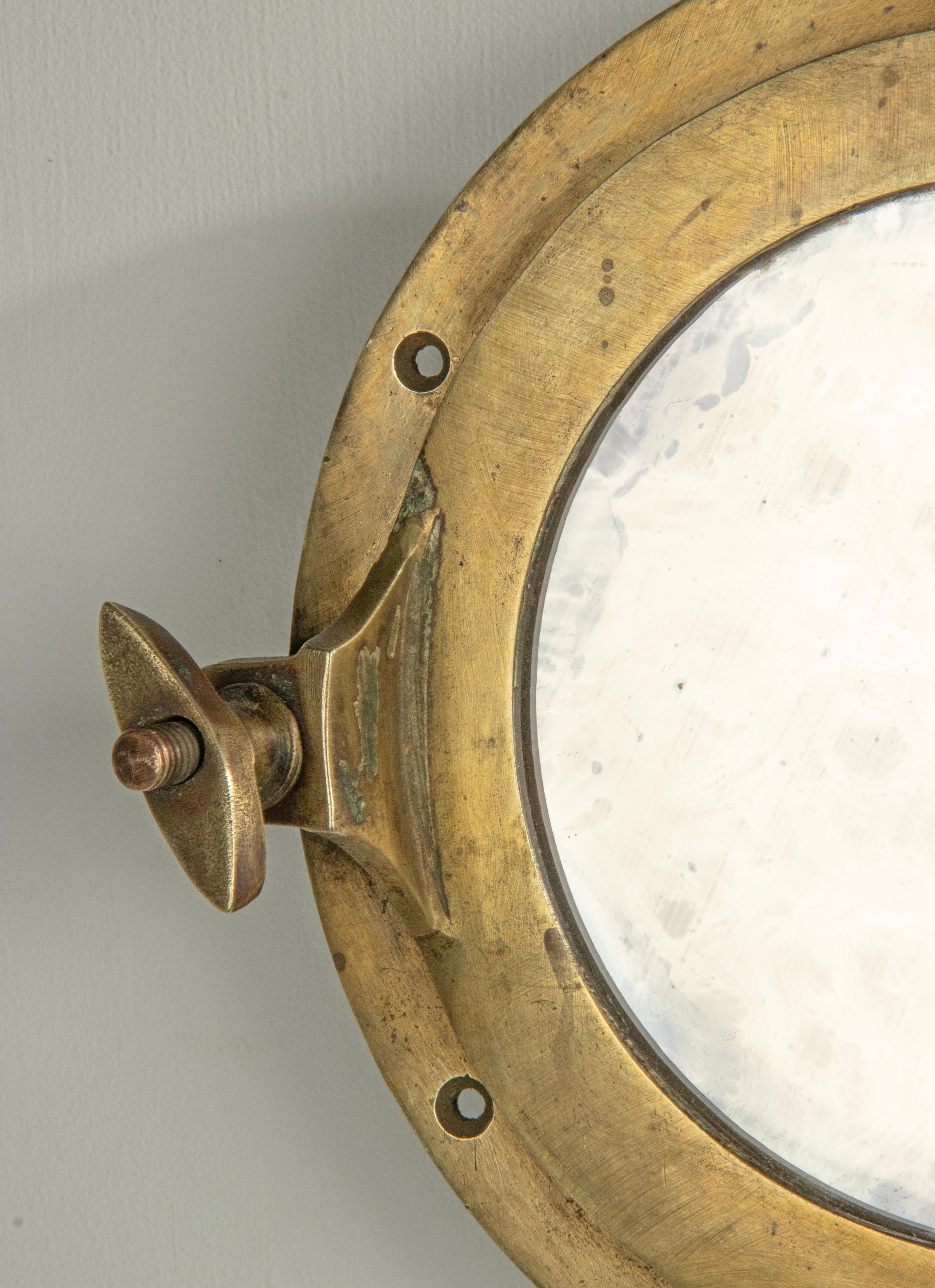 A small antique ship porthole wall mirror made of brass. A mirror glass has been placed inside. The whole has a beautiful old patina. The original glass is removed and replaced by a mirror glass. Some wear stains on the mirror glass. This maritime