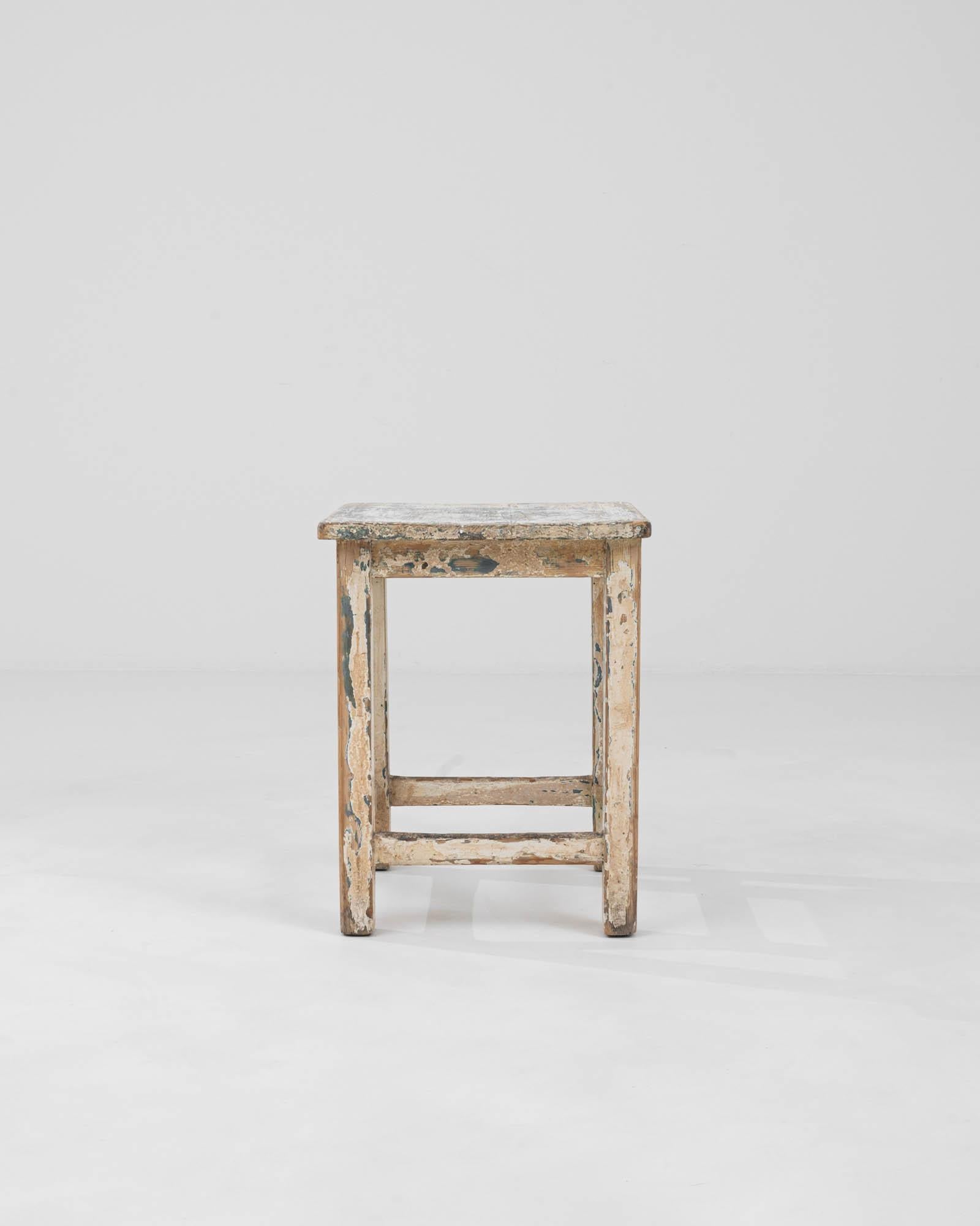 This Early 20th Century Central European stool is a testament to the timeless beauty of patinated wood, offering a sense of history and nostalgia. Its robust wooden frame, displaying layers of flaking paint in muted tones, tells a rich story of its