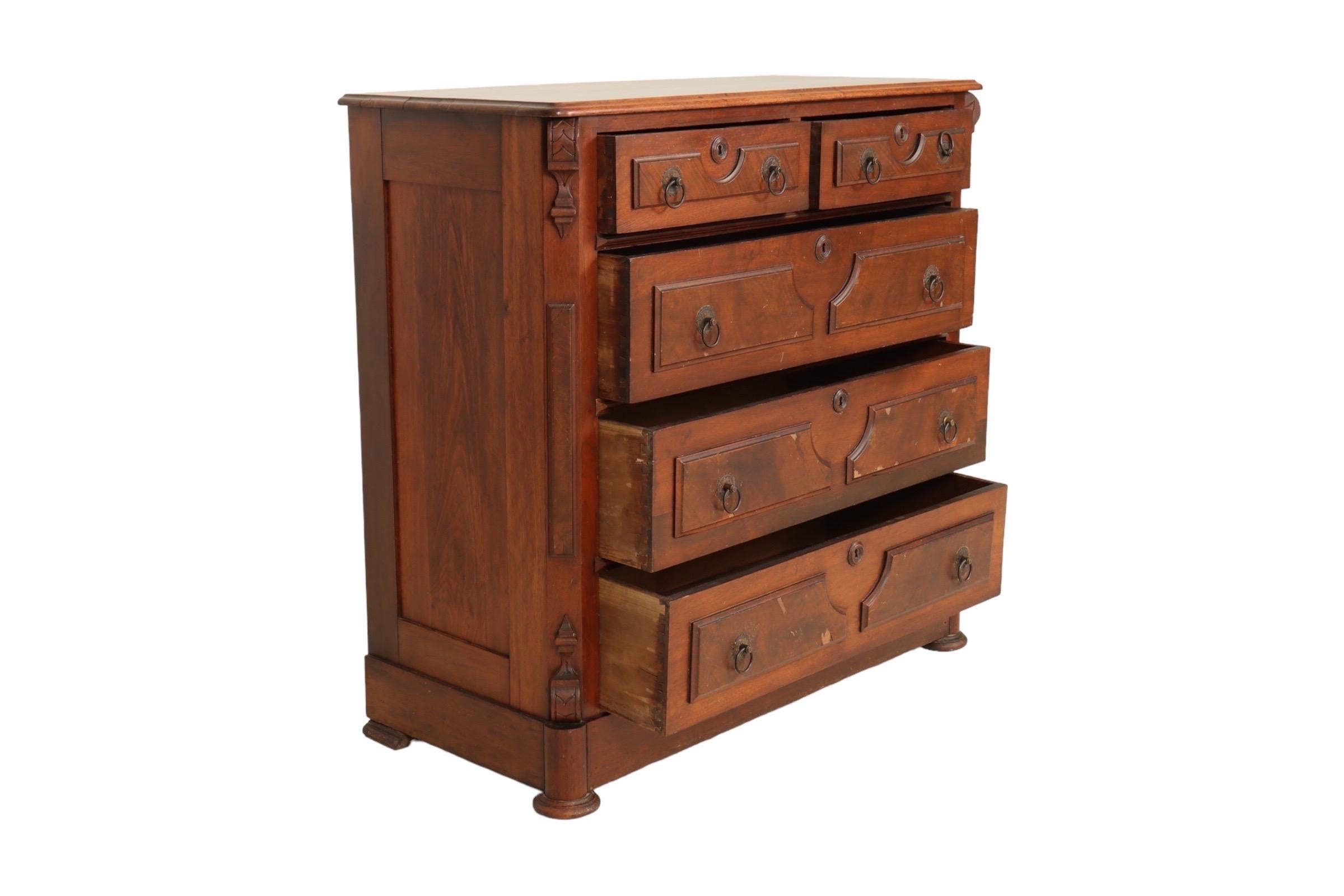 An early 20th century chest of drawers made of walnut. Two small drawers above three long drawers each have symmetrical wooden veneer panels with beveled surrounds and brass drop hoop handles on decorative round back plates. The Eastlake style