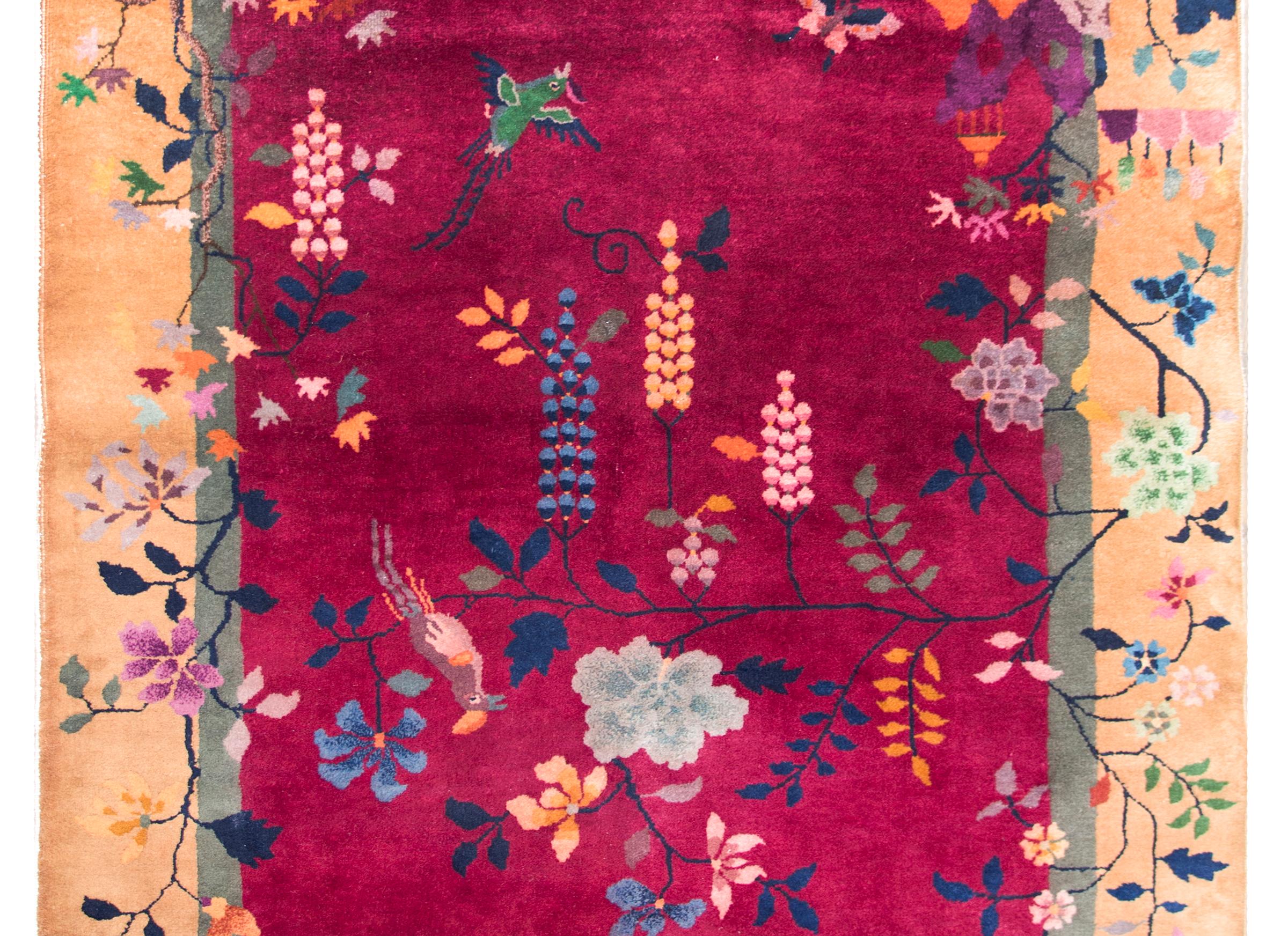 A beautiful early 20th century Chinese Art Deco rug with a fuchsia background, champagne colored border, and overlaid with myriad auspicious flowers including peonies, chrysanthemums, and branch with birds hanging over a pagoda in a garden landscape.