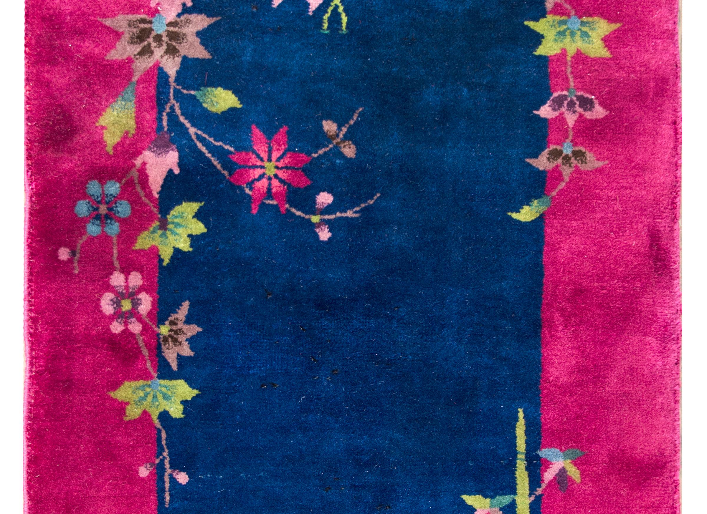 A wonderful early 20th century Chinese Art Deco rug with a deep indigo field surrounded by a brilliant fuchsia border, and all overlaid with multi-colored auspicious flowers including chrysanthemums, peonies, lotus, and cherry blossoms.  