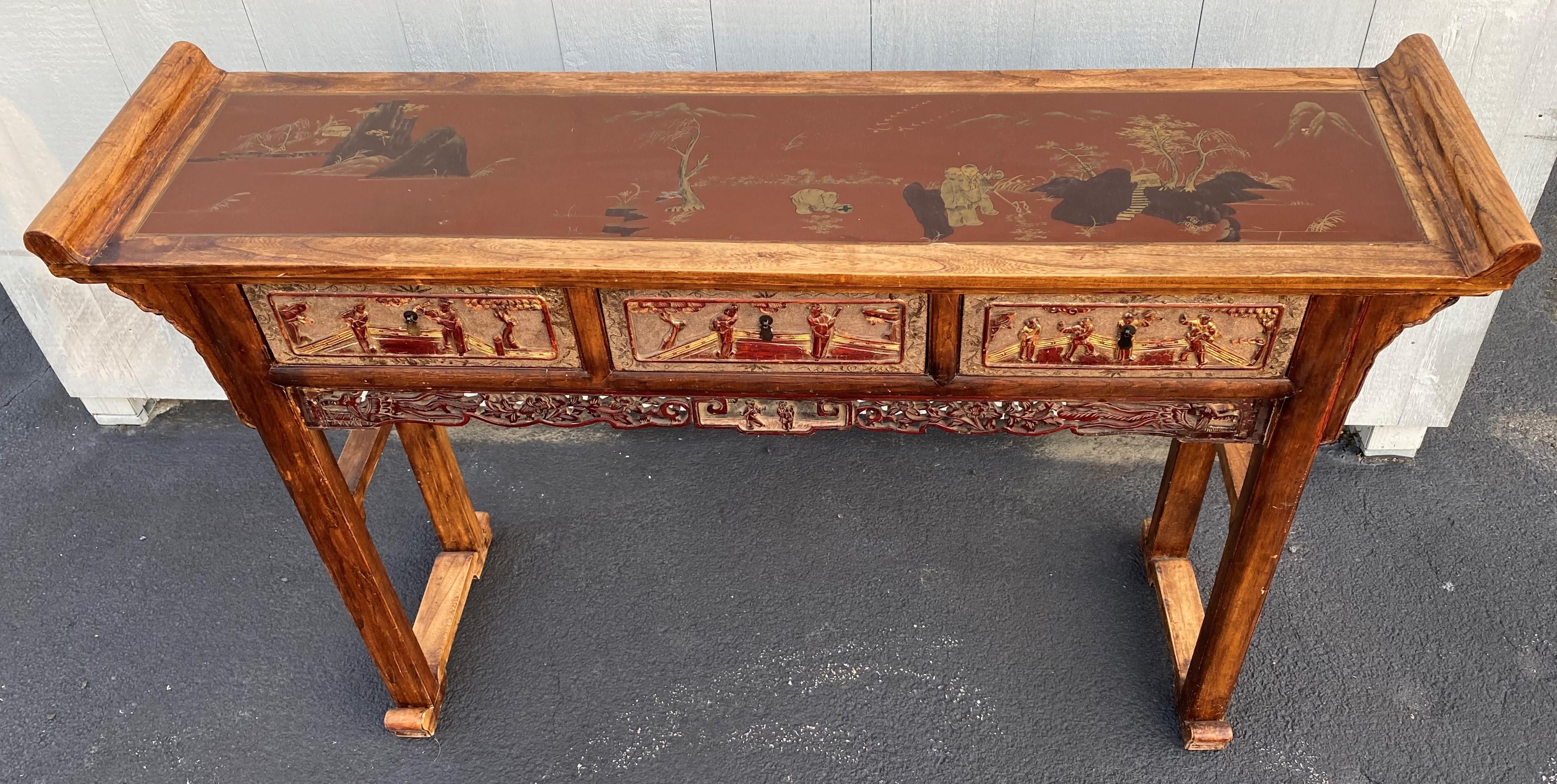 A finely carved polychrome three-drawer hardwood altar table with relief carved genre scenes on each drawer front and the top of each end, as well as painted scenes with landscapes and figures on an oxblood background the top, as well as a