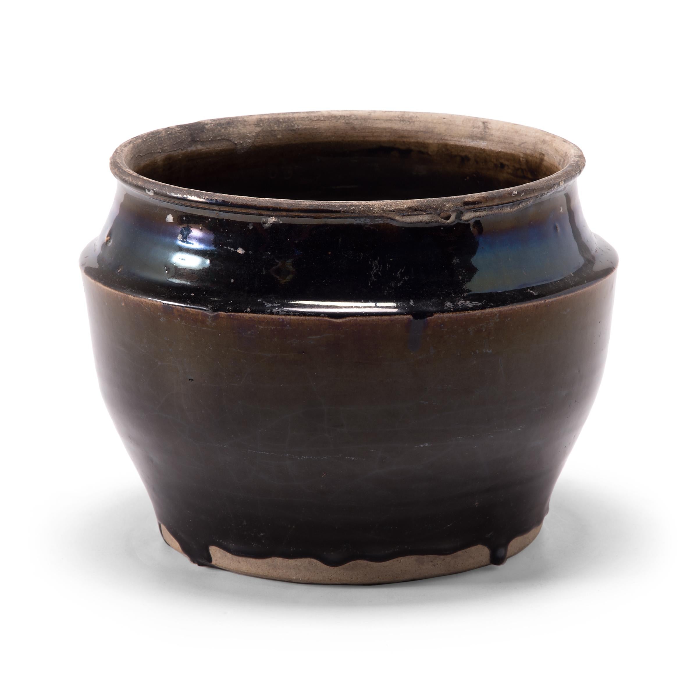 As though slicked with oil, a thick blue-black glaze spills over this squat kitchen vessel, pooling at the angular shoulders for an iridescent shimmer. The early 20th century ceramic basin was once used for pickling foods in a Qing-dynasty kitchen,