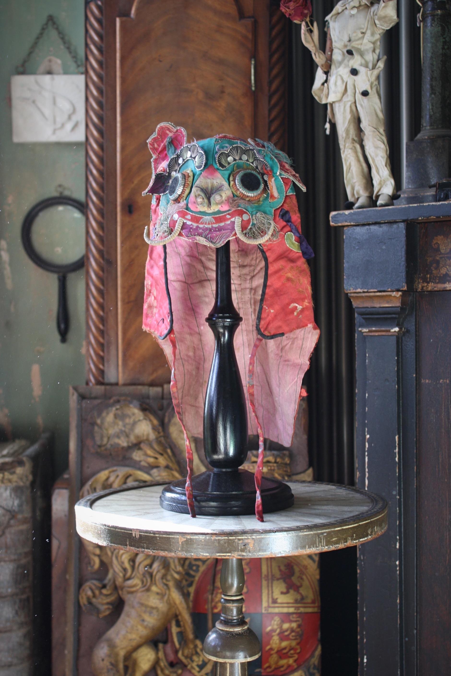 A Chinese silk hat from the late Qing period, displayed on a purpose made antique style ebonised wig stand.

These head dressers traditionally worn by children and often displayed mythical creatures, formed from embroidered features with a playful