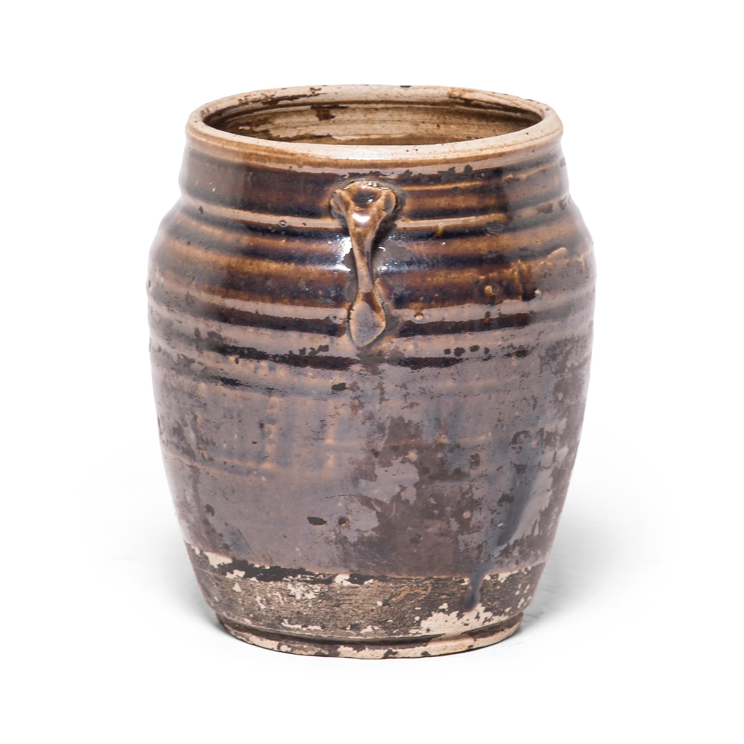 A thin brown glaze clings to the high shoulders of this petite jar, pooling between subtle ridges to create a pattern of light and dark across the vessel. Crafted in the late 19th century, the wide-mouth jar with strap handles was once used to store