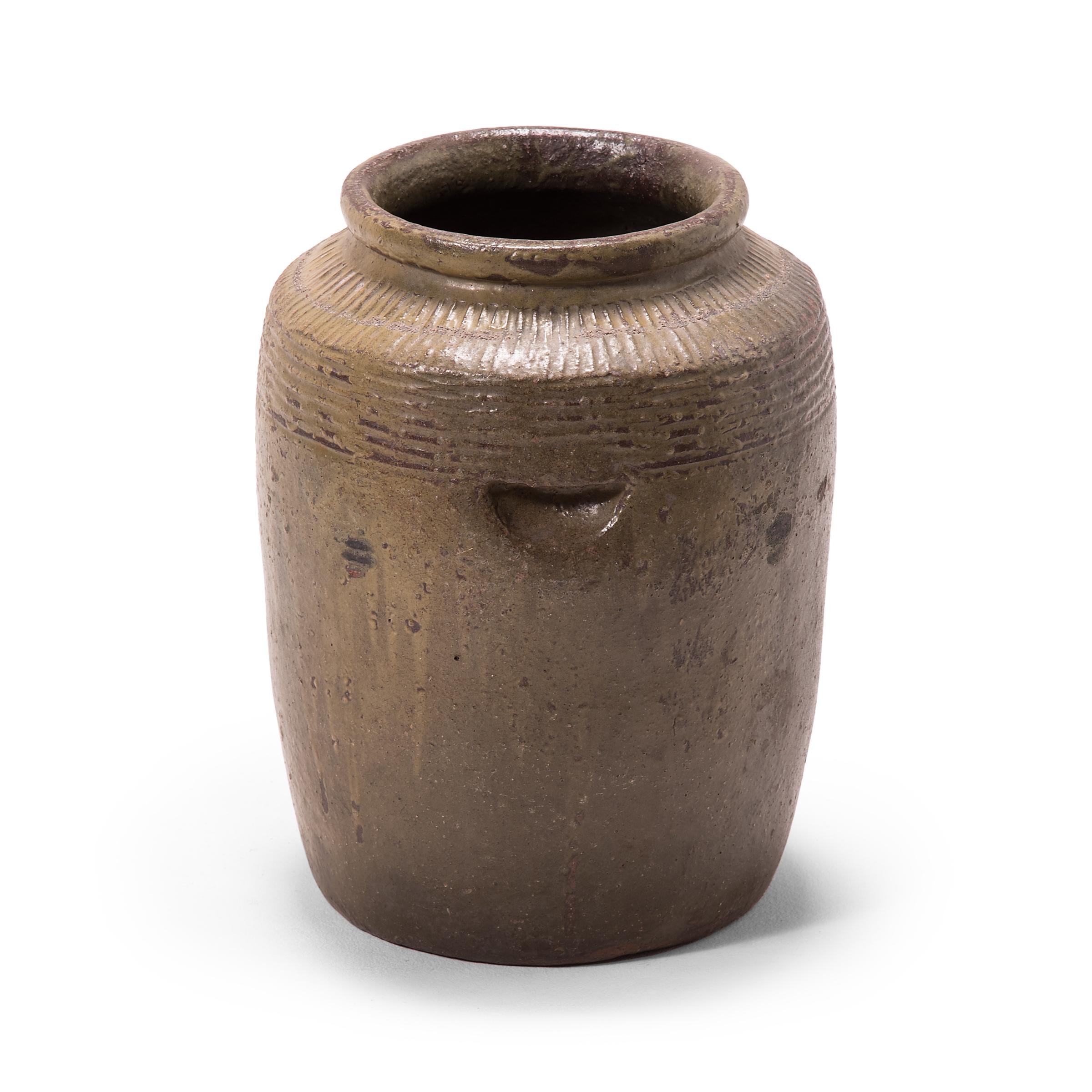 Originally used for pickling foods, this early 20th century ceramic jar is coated inside and out with a warm grey glaze. Decorative ridges patterning the jar's high shoulders manipulate the glaze to pool irregularly across the surface. The jar is