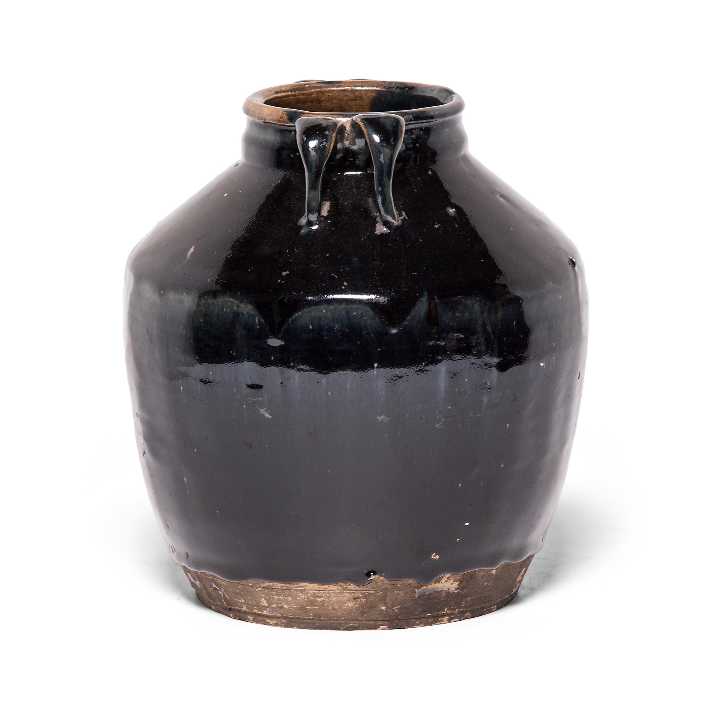 As though slicked with oil, a thick blue-black glaze spills over the high shoulders and arched strap handles of this tapered kitchen vessel. The 19th century ceramic jar was once used to prepare and store soy sauce in a Qing-dynasty kitchen, as