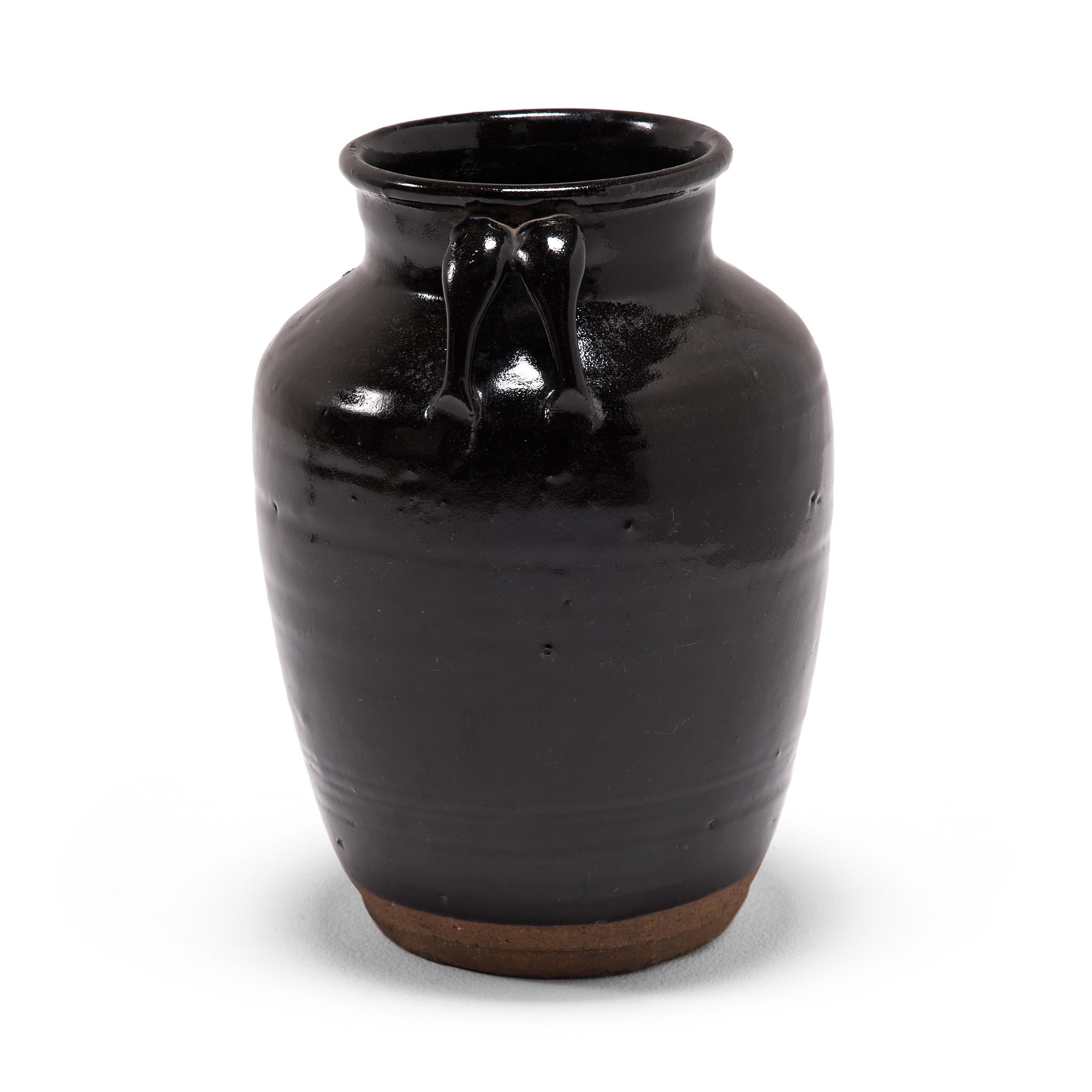 As though slicked with oil, an inky black glaze drenches the high shoulders and arched double strap handles of this tapered kitchen vessel. The 19th century ceramic jar was once used to prepare and store soy sauce in a Qing-dynasty kitchen, as