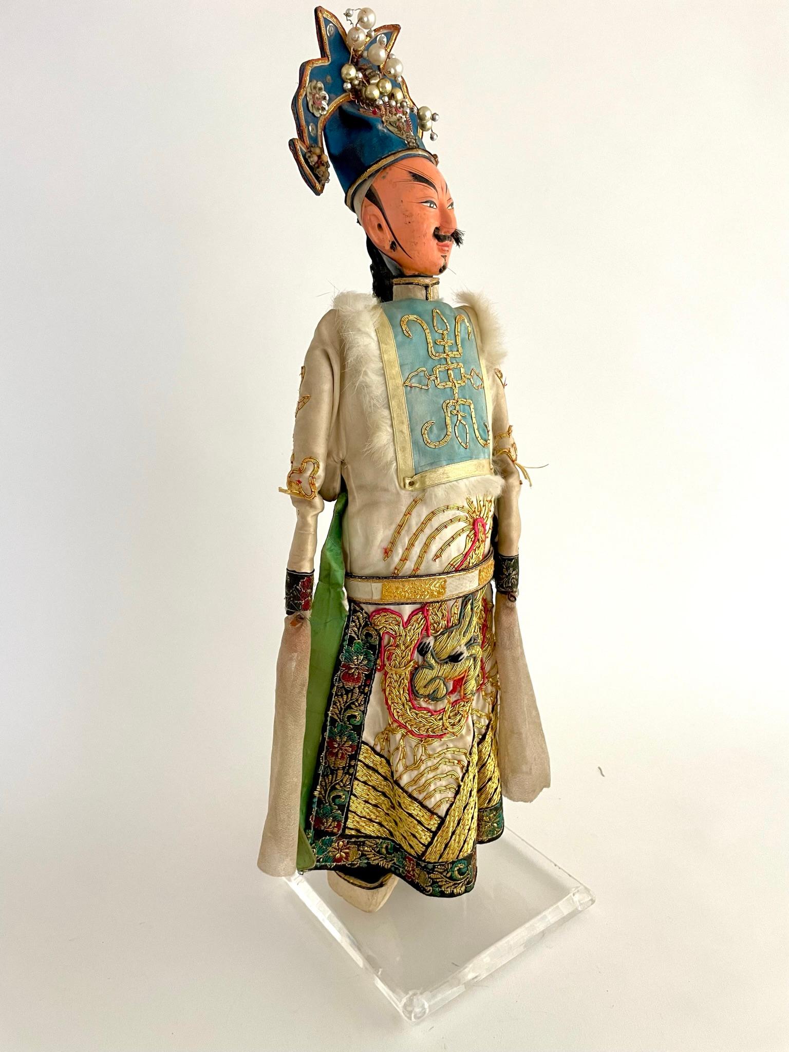 Diese chinesische Opernmarionette ist in ein elegantes, besticktes Drachengewand aus Seide gekleidet. Der Puppenkopf ist aus bemalter Keramik. Der kunstvolle Kopfschmuck ist mit Perlen, Seide und Metallornamenten verziert. Diese Puppen wurden zu