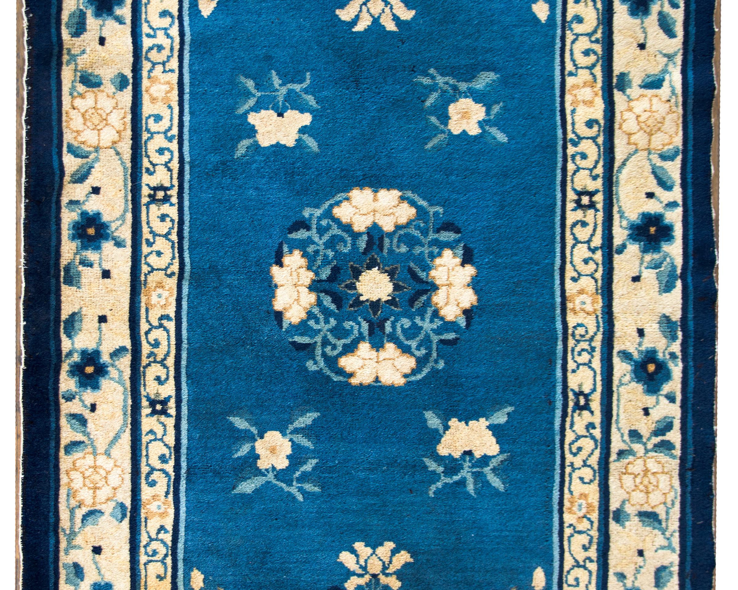 A wonderful early 20th century Chinese Peking rug with a central peony medallion living amidst a field of more peonies, chrysanthemums, and lotus flowers, all woven in cream, brown, and dark indigo set against a light indigo background.  The border