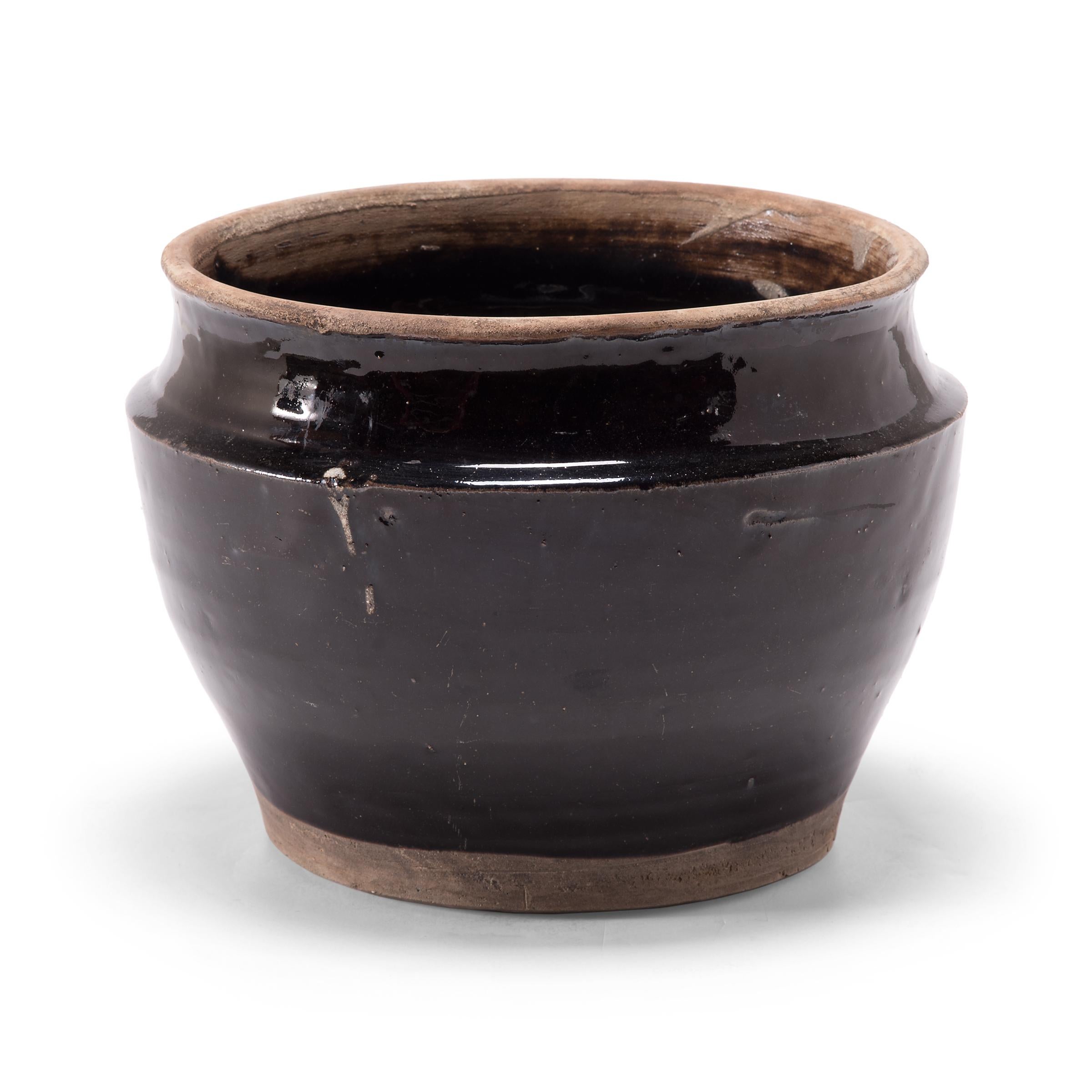 A dark glaze cloaks the high, angular shoulders and tapered body of this squat kitchen vessel, save for the unfinished lip and base. The early 20th century ceramic basin was once used for pickling foods in a Qing-dynasty kitchen, as evidenced by its