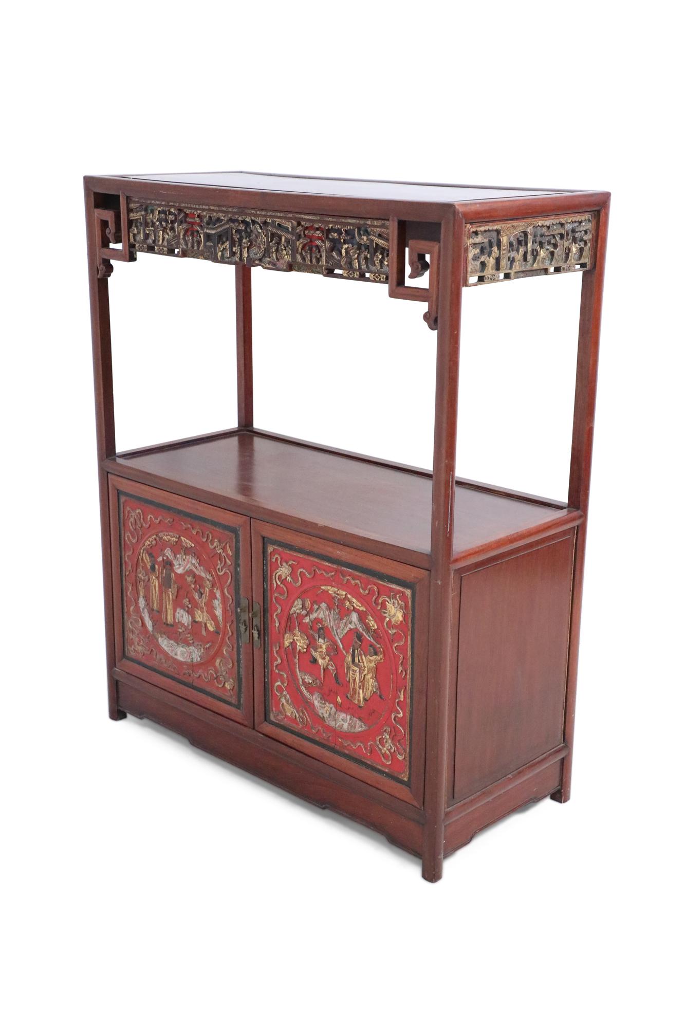 Chinese (early 20th Century) red wooden display cabinet with an open main shelf framed above a cabinet, and intricate gold figurative scenes carved into both the doors, and a top border.