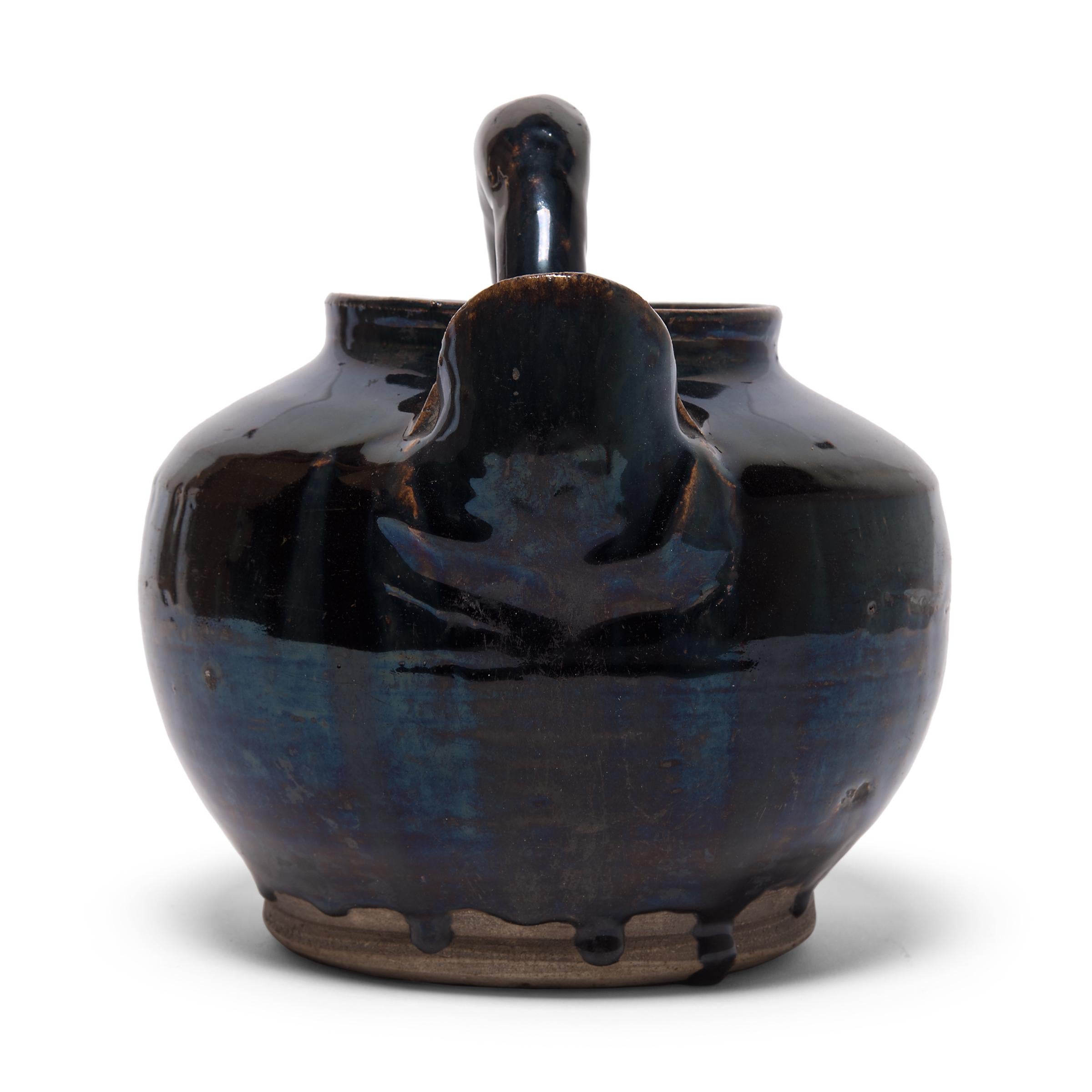 A dark blue-black glaze coats the round body of this squat kitchen vessel, dripping down its sides with subtle iridescence. The early 20th century spouted vessel was once used for storing wine or soy sauce in a Qing-dynasty kitchen, as evidenced by