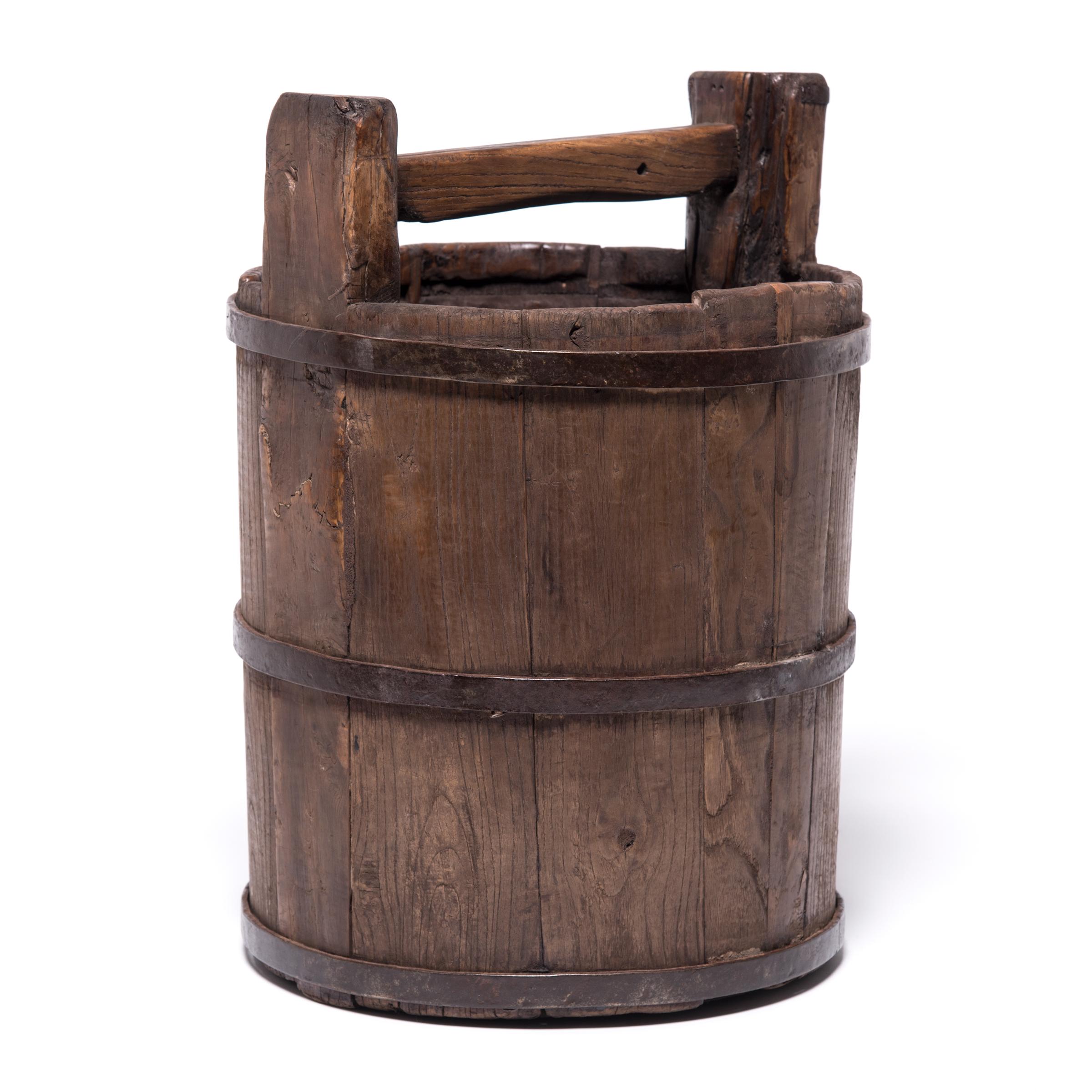 Beautifully aged with a warm patina, this Qing-dynasty bucket draws attention to the ingenuity of its manufacture, consisting of staves of cypress bound with iron rings. Conjuring up images of the water-bearing figures illustrated in painted