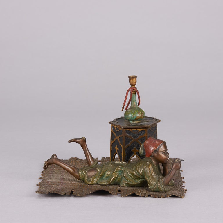 An excellent early 20th Century Austrian bronze study of a young Arab boy wearing a fez and holding a cigar whilst reclining on a straw rug next to an Orientalist table, hinged to open as a working inkwell with original fitment. The surface of the