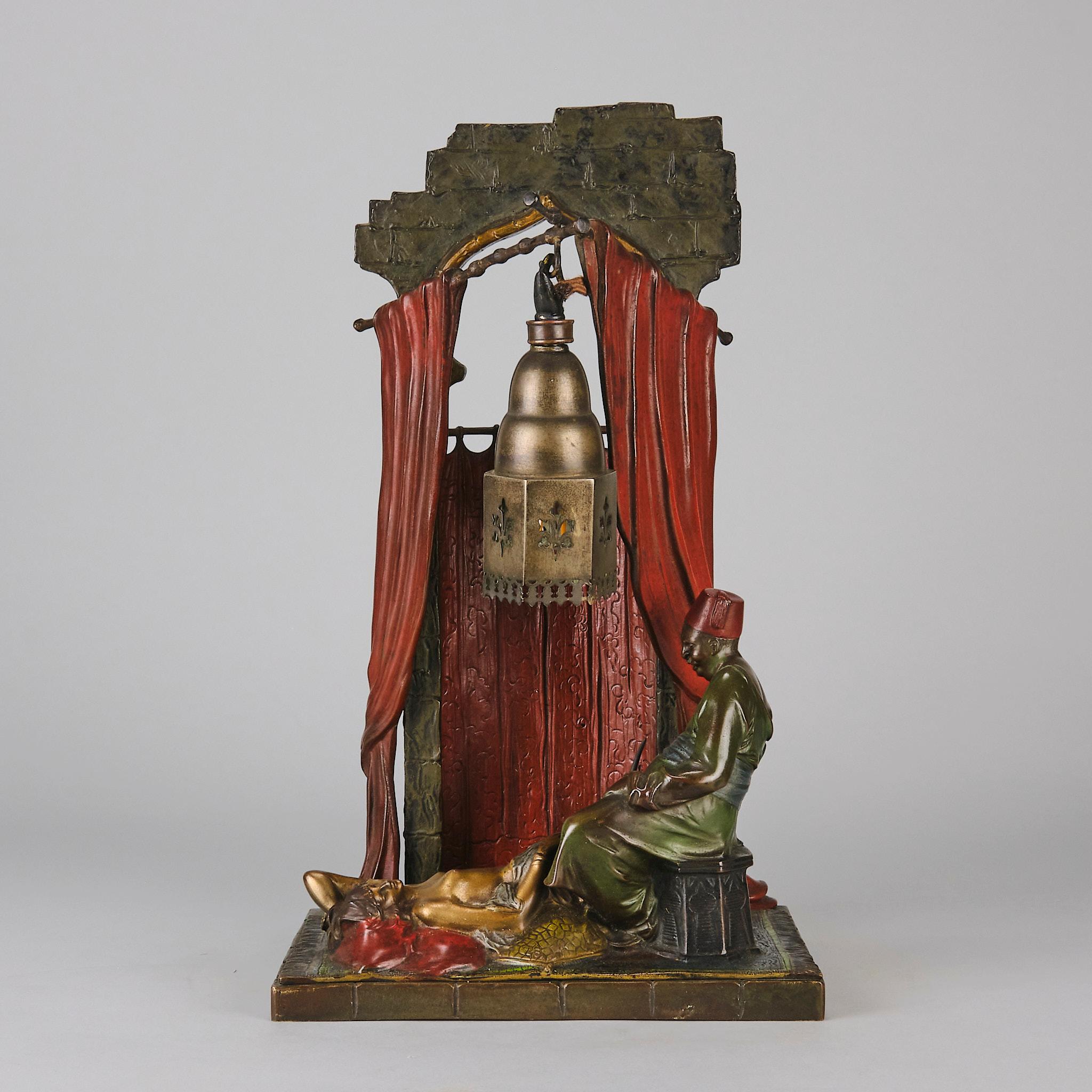 An erotically charged early 20th century Art Deco cold painted bronze lamp modelled with a harem beauty relaxing on a rug in front of an Arab man with an architectural background with suspended lantern containing a light. The whole raised on a