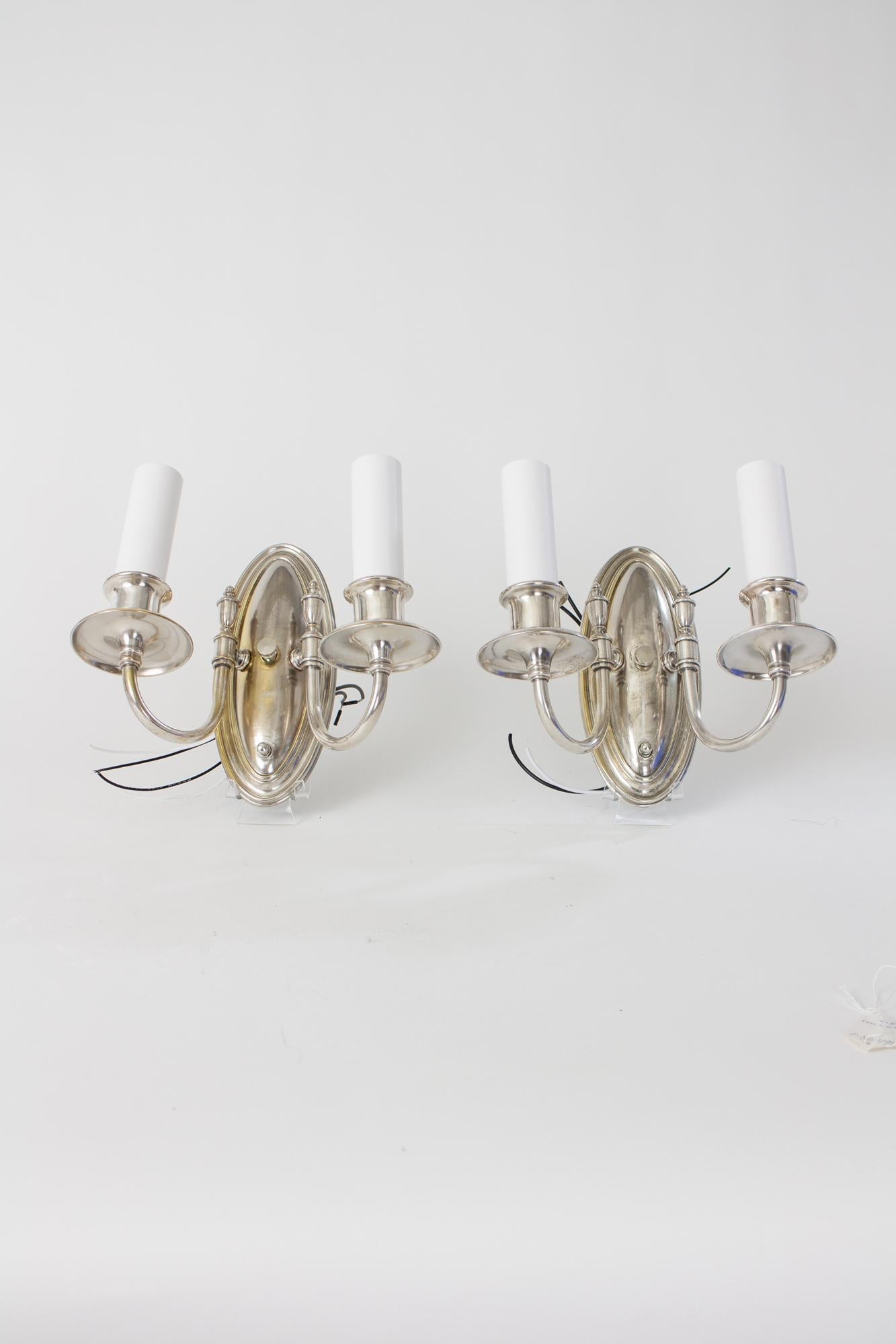 Early 20th century double arm silver sconces. Neoclassical style. Oval backplate with delicate arms that reach up to two lights. Urn shaped ornament on arms, White Candlecovers. Restored and rewired. Complete and ready for installation. Worn antique