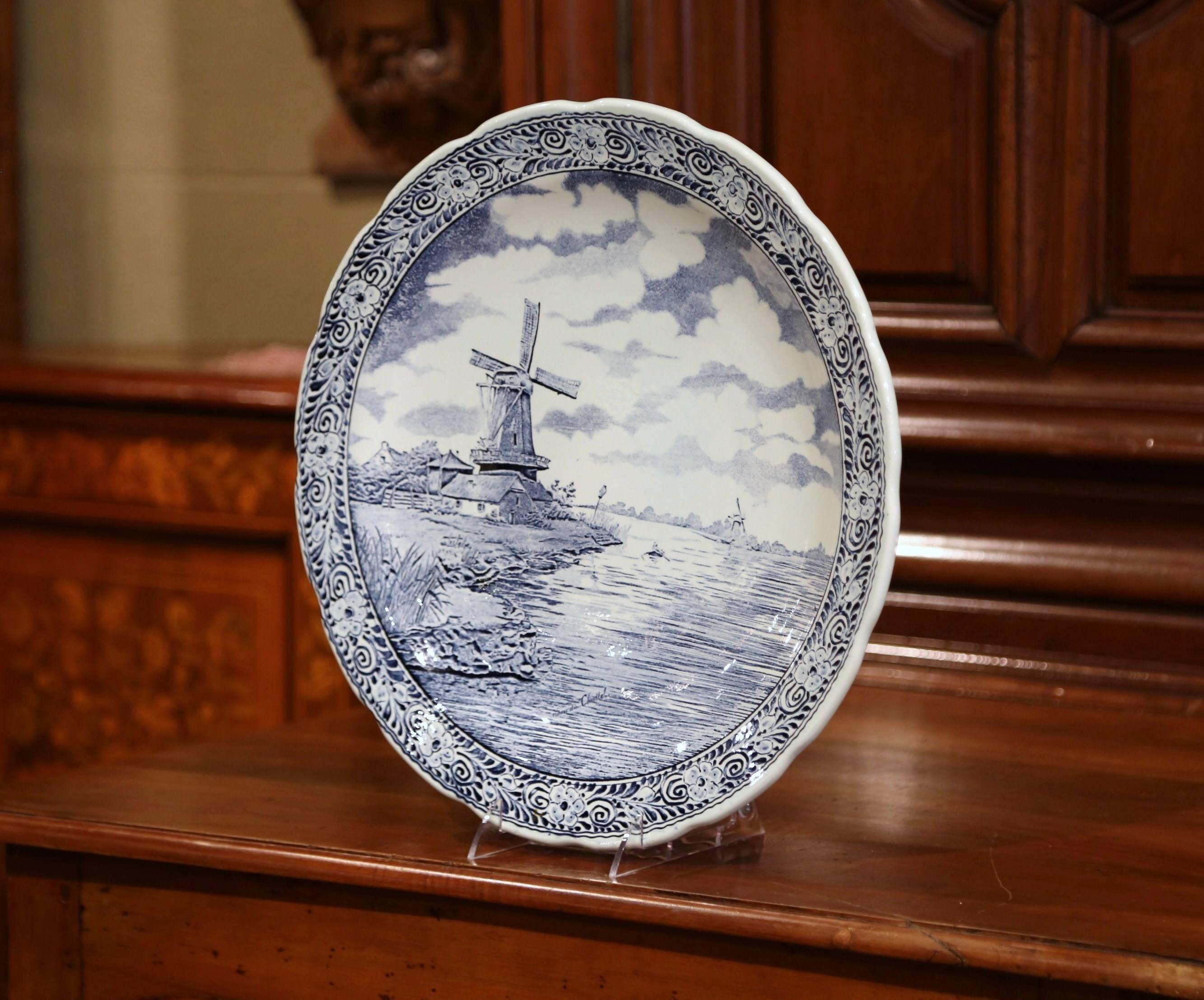 This large antique platter was crafted in Holland, circa 1920. The large, wall hanging plate depicts a lake scene with a traditional Dutch windmill in the Classic blue and white palette. The ceramic piece has a flower border and features a farm