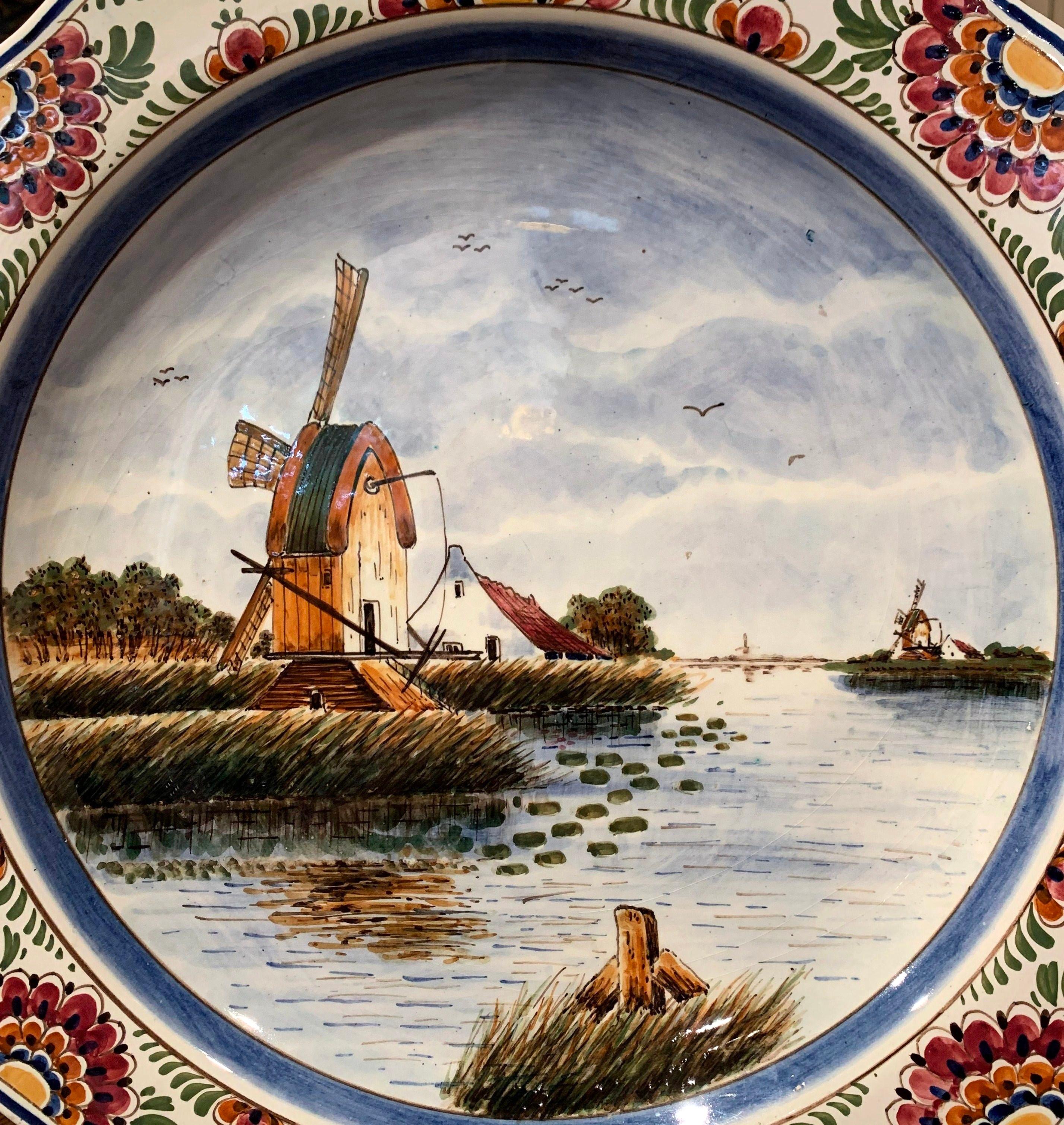 This large colorful charger was crafted in Holland, circa 1930, round in shape, the faience dish features traditional hand painted delft decor, including windmill, farmhouse and lake, it is further embellished with floral decor around the rim. The