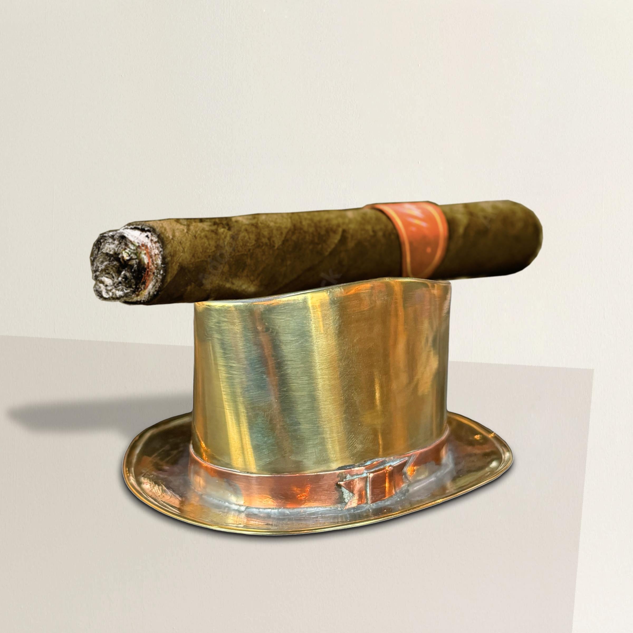 This charming early 20th-century English Edwardian brass top hat cigar holder is a delightful piece for any cigar aficionado or collector of whimsical decor. Hand-crafted in the shape of a classic top hat, the gleaming brass body features a dimpled