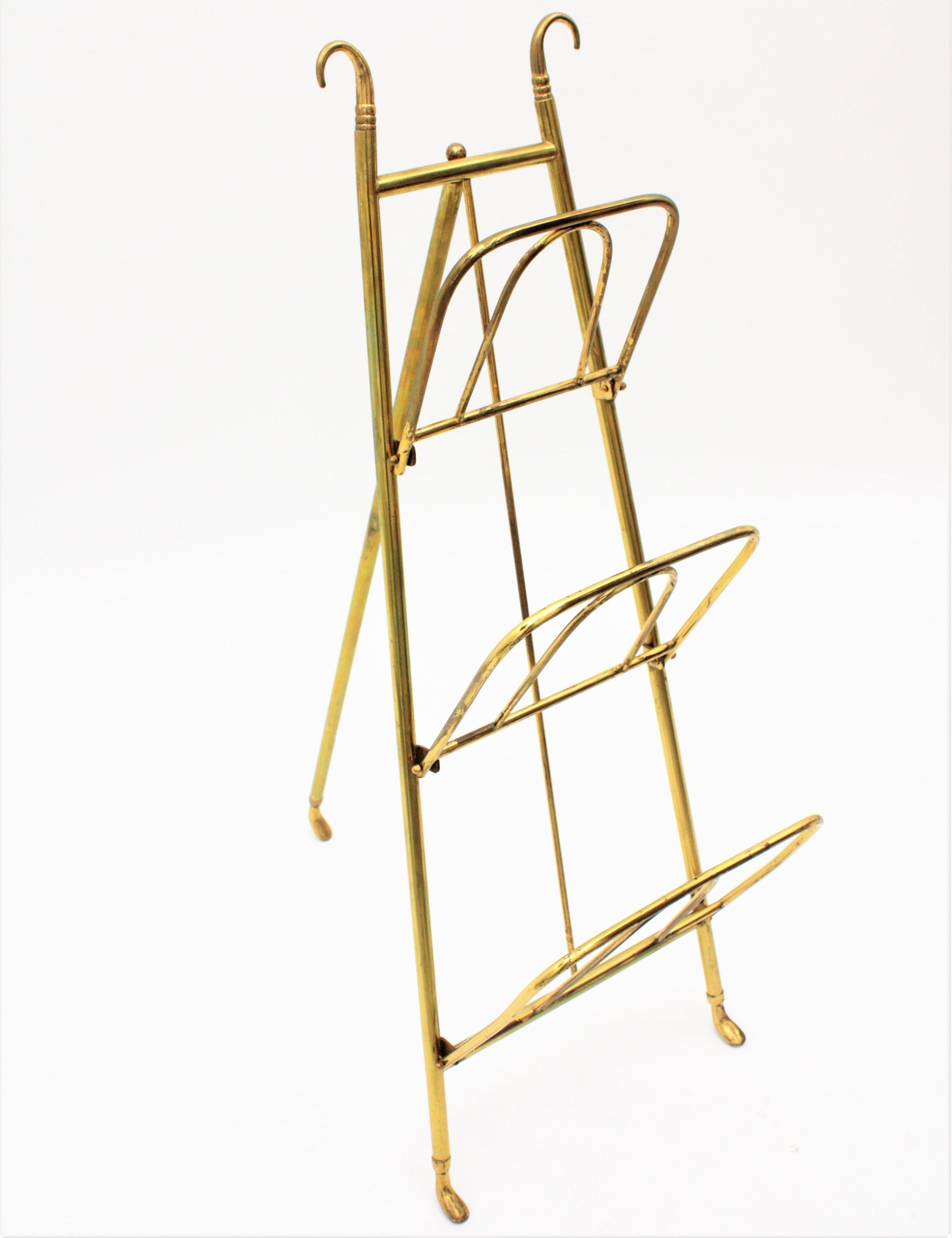 Edwardian polished brass tripod magazine rack with three tiers. It has stylish feet endings at the bottom and hooks endings at the top. 
England, 1910-1920.
Use it as a magazine, book or newspapers rack, easel or towels valet in a powder room. Easy