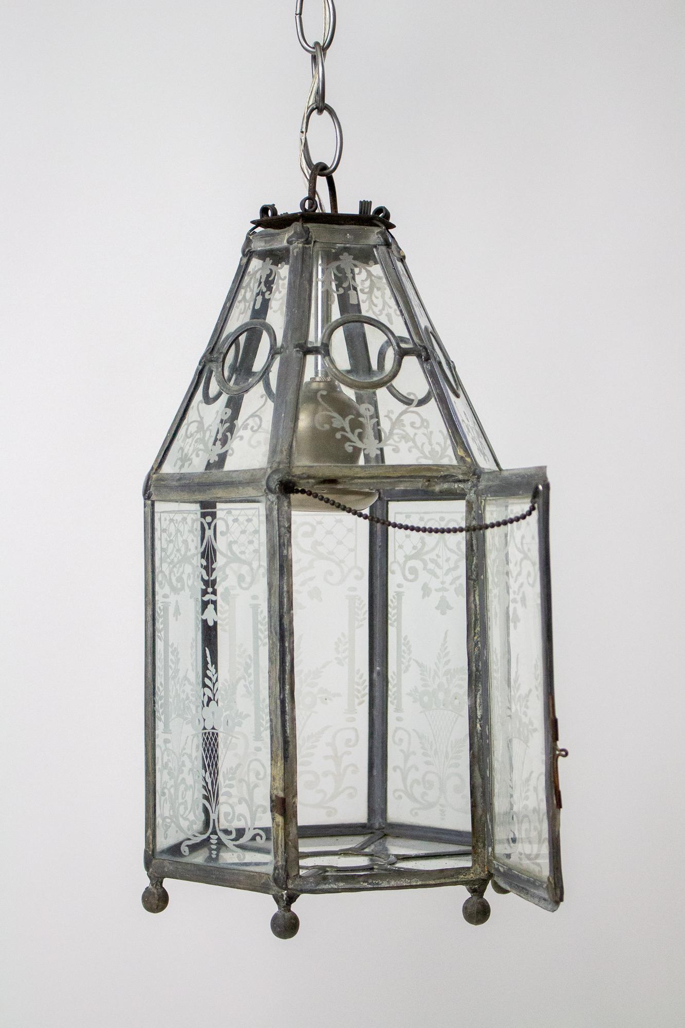 A hexagonal leaded glass lantern. The glass panels are wheel engraved with a curved foliate flower basket design. The top panels slant to the top and have clear round glass pieces. The bottom is flat with a circular center panel and six trapezoidal
