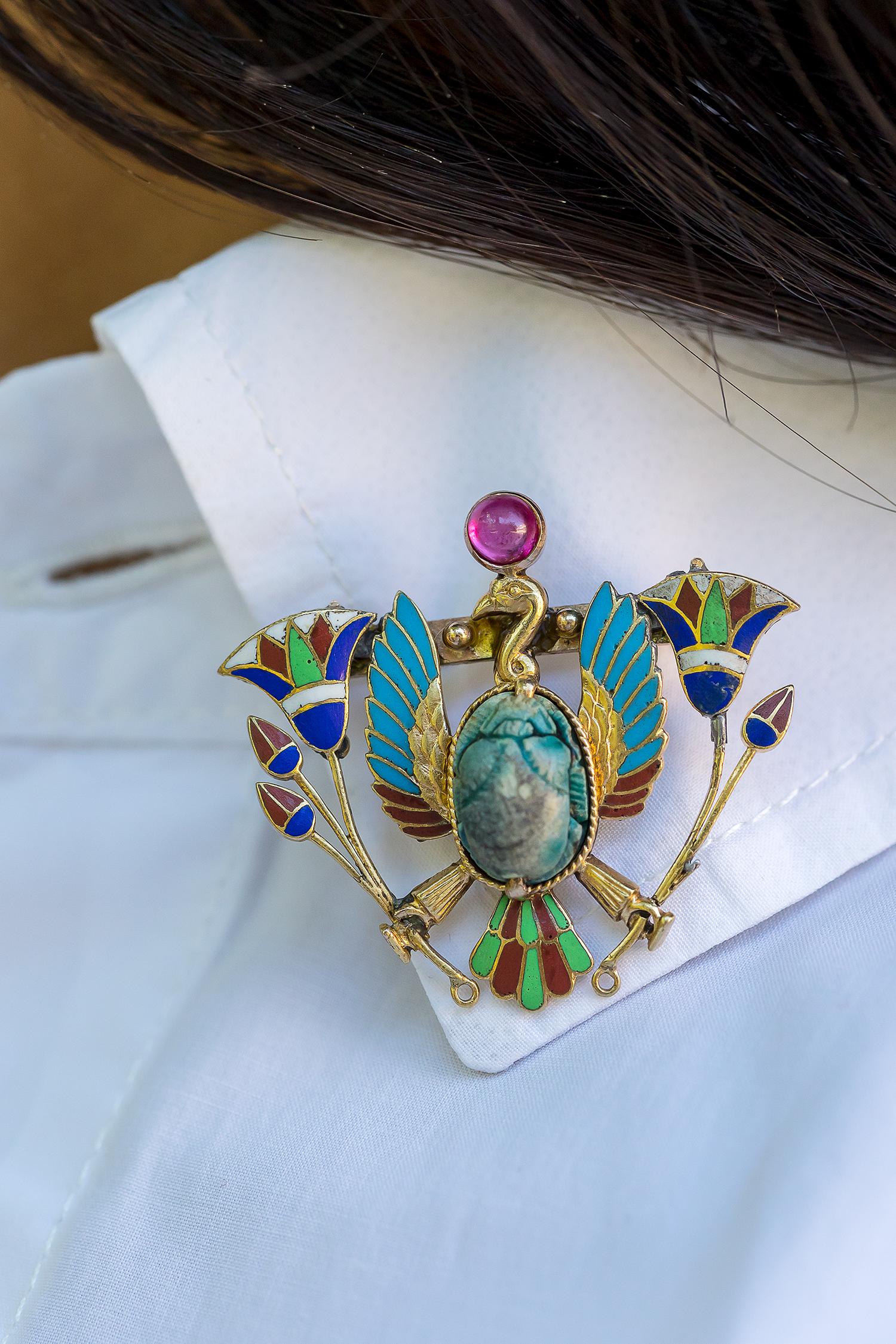 An early 20th century Egyptian Revival gold brooch, depicting a vulture centred with a faïence scarab, clutching lotus pods and surmounted with a cabochon ruby, polychrome enamel decoration overall and engraved to reverse, 3.5cm wide. One arrow