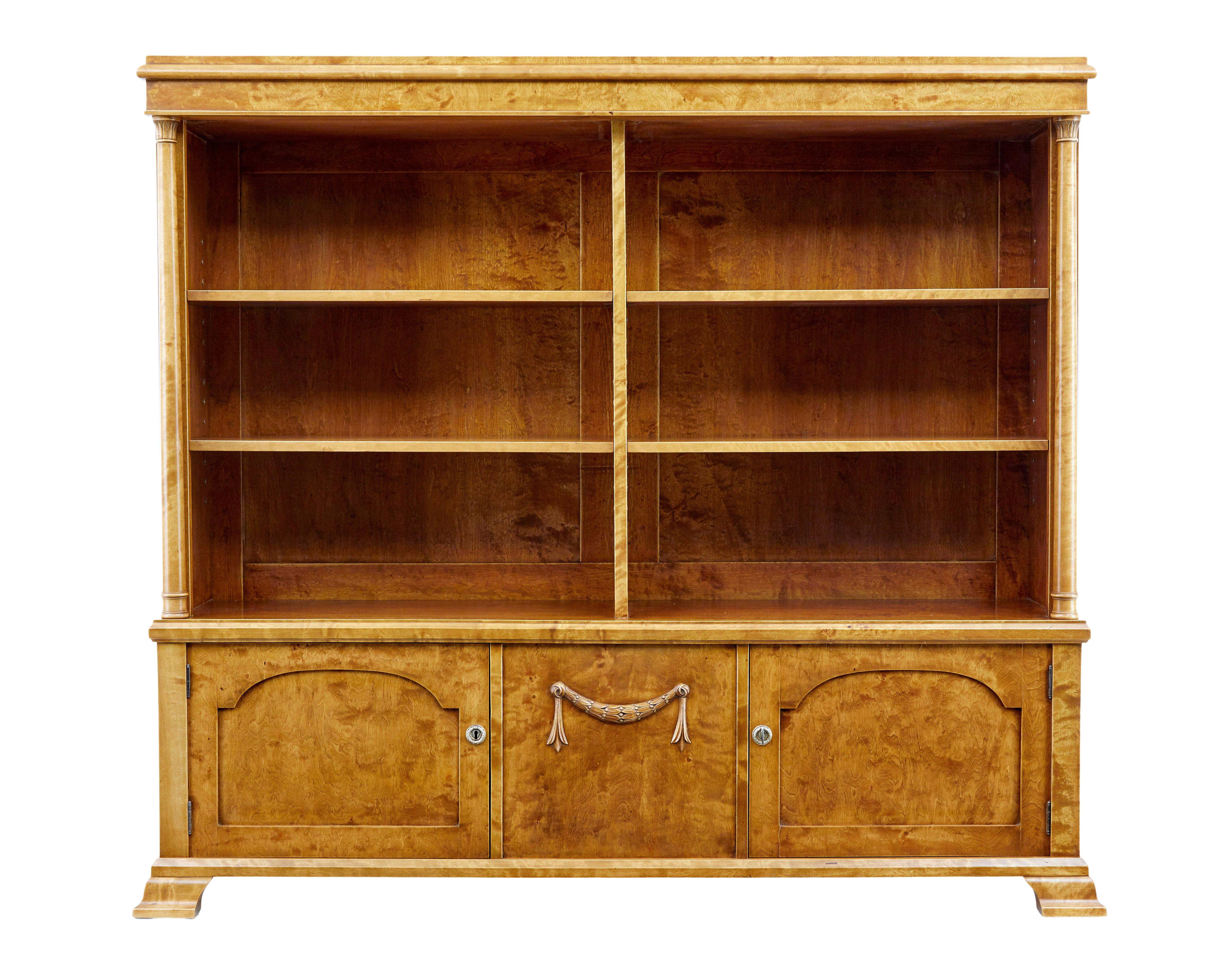 Beautiful rich birch open bookcase, circa 1910.

Four adjustable shelves with a central partition, below a central panel with carved applied swag, flanked either side a single door cupboard.

Turned columns run up both front sides and link to the