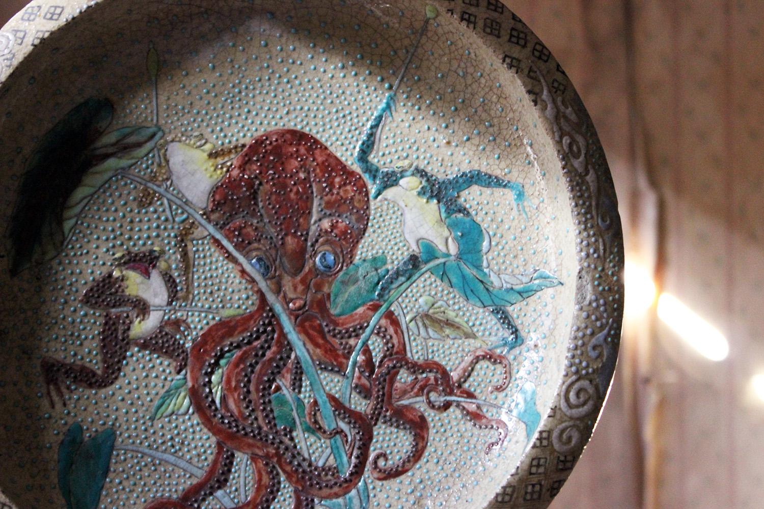 The Japanese stoneware satsuma charger, painted in bright enamels and a slip glaze depicting a red octopus or kraken being hunted by three frogs with pampas grass as spears, the octopus clutching a lily, to a spotted ground and patterned rim with