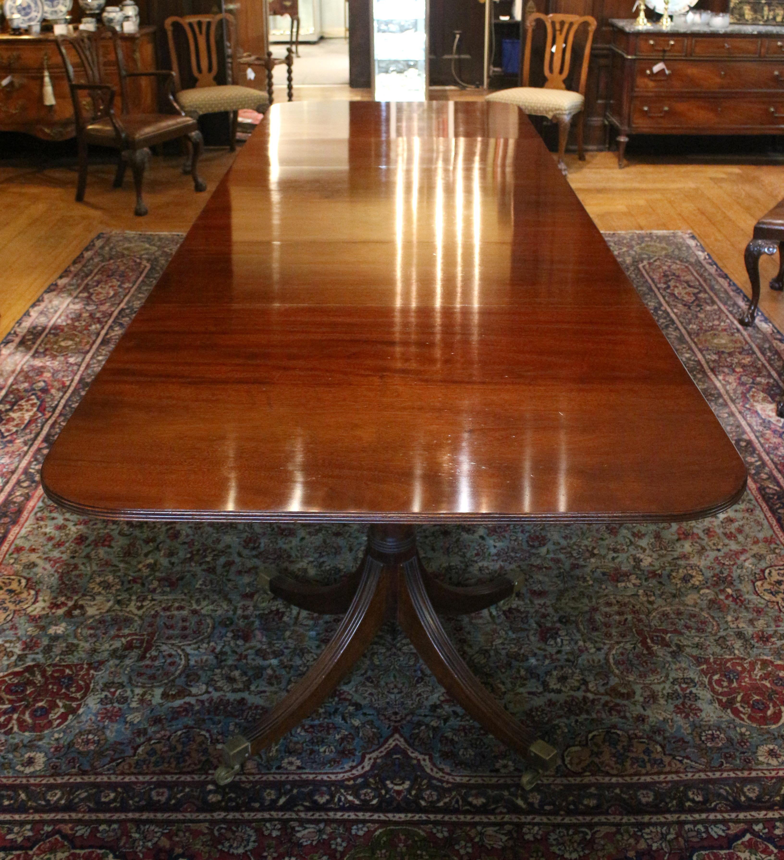 Mesa de comedor banquete de triple pedestal de estilo georgiano de principios del siglo XX, inglesa. 2 hojas. Elevadas sobre bases cuadrúpedas torneadas en anillo, las patas estriadas terminan en cazoletas cuadradas de latón y ruedecillas. Maderas